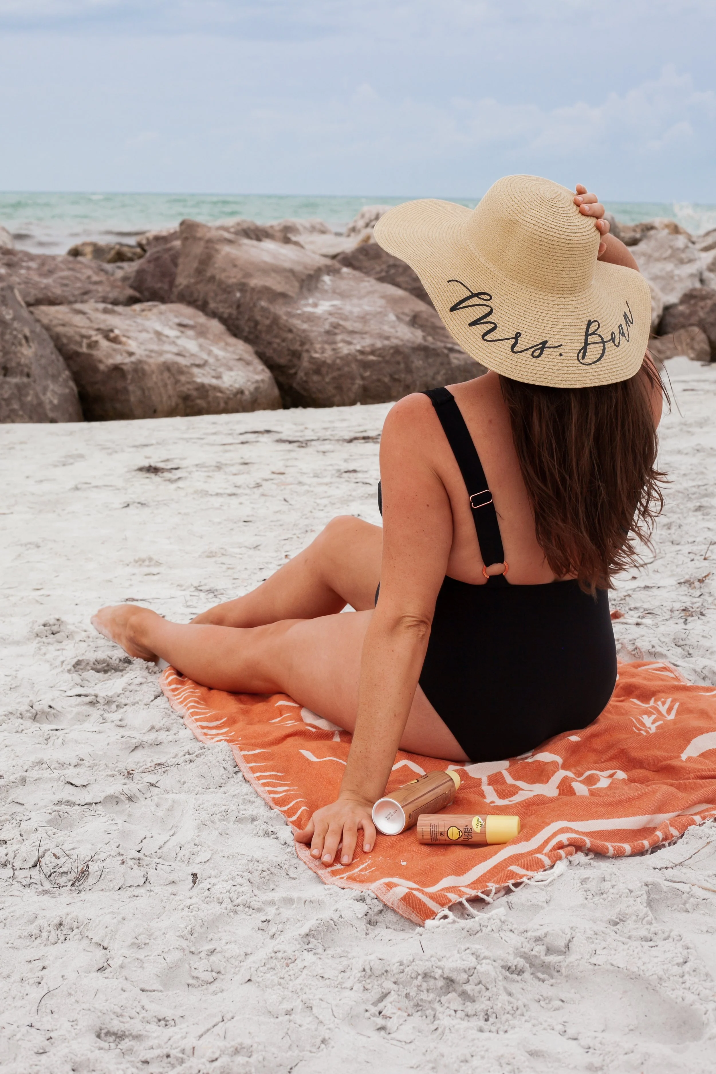 Woman sitting on an orange towel on a sandy beach, wearing a black swimsuit and a large straw hat with 'Mrs. Beau' written on it, with some lotion bottles nearby, rocks, and the ocean in the background.