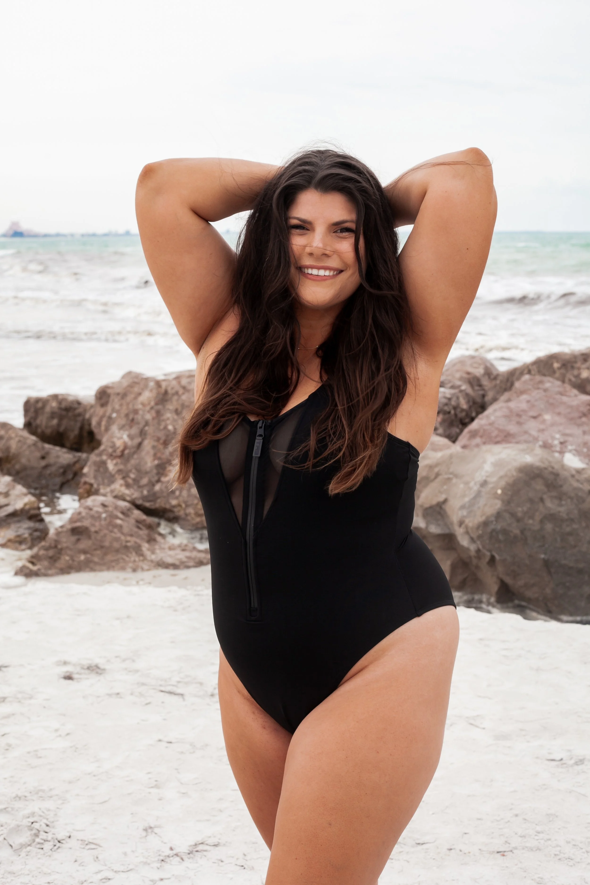 A woman in a black swimsuit standing on a sandy beach with rocks and the ocean in the background, smiling with her arms raised behind her head.