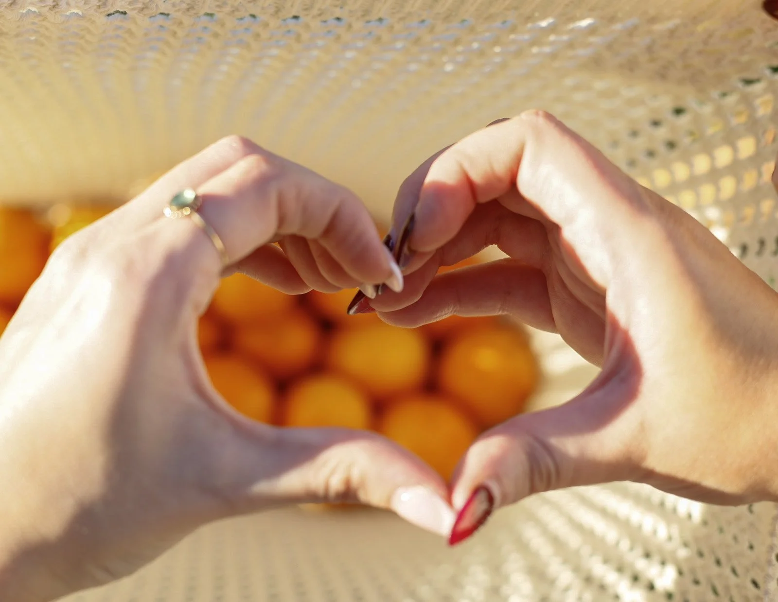 Two hands forming a heart shape in front of a basket of oranges.
