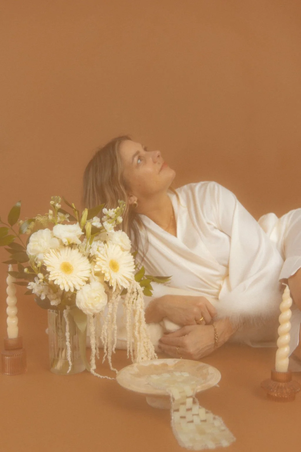 A woman with light brown hair sitting at a table with a bouquet of white flowers, looking up, against a plain brown background.
