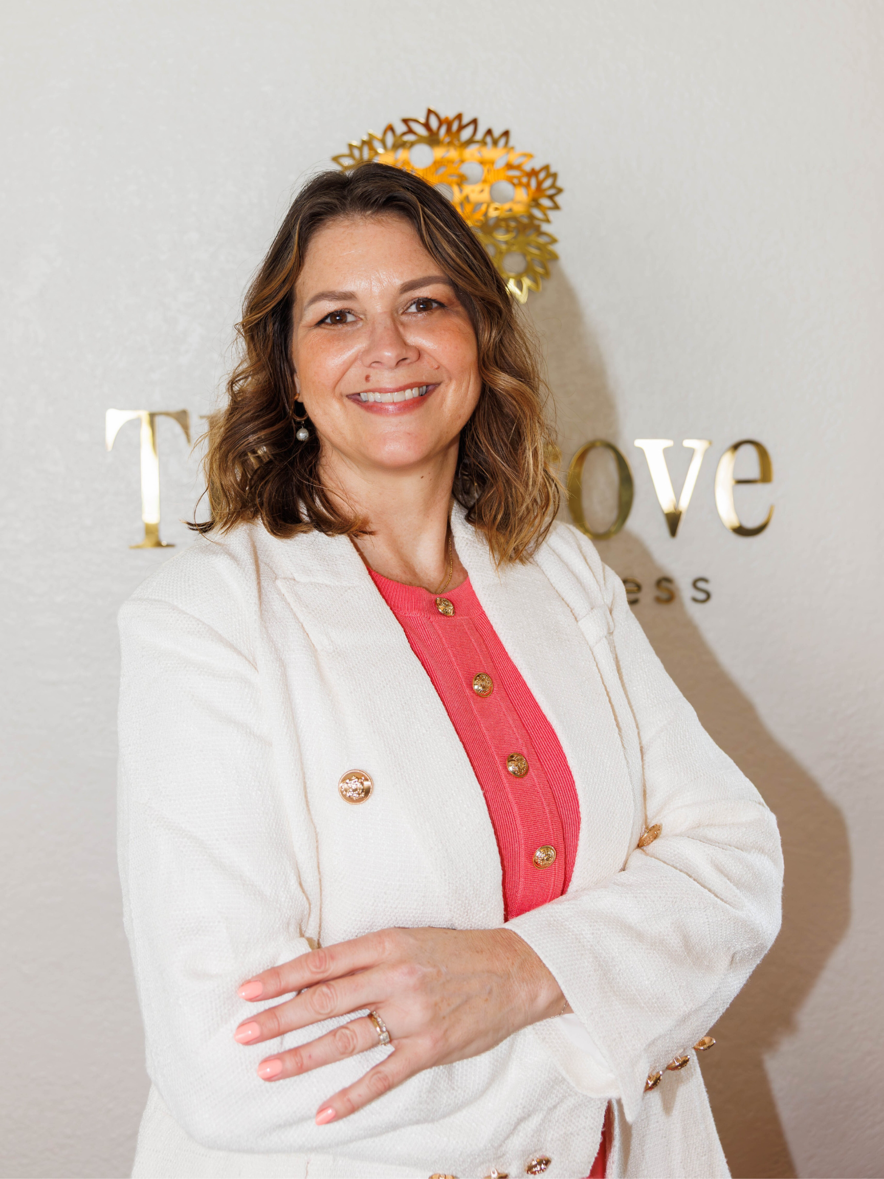 A smiling woman with shoulder-length wavy brown hair, wearing a white blazer with gold buttons over a pink top, standing in front of a wall with a decorative gold and white sign that reads 'T Love'.