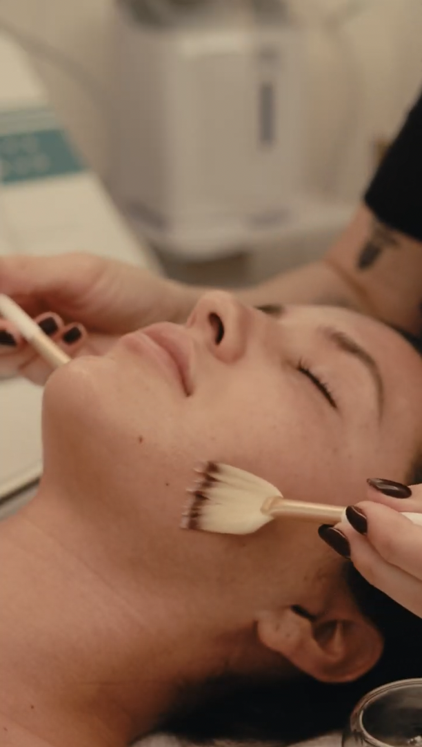 A person receiving a facial treatment, with closed eyes, as a skincare professional applies a product using a fan brush on their cheek.