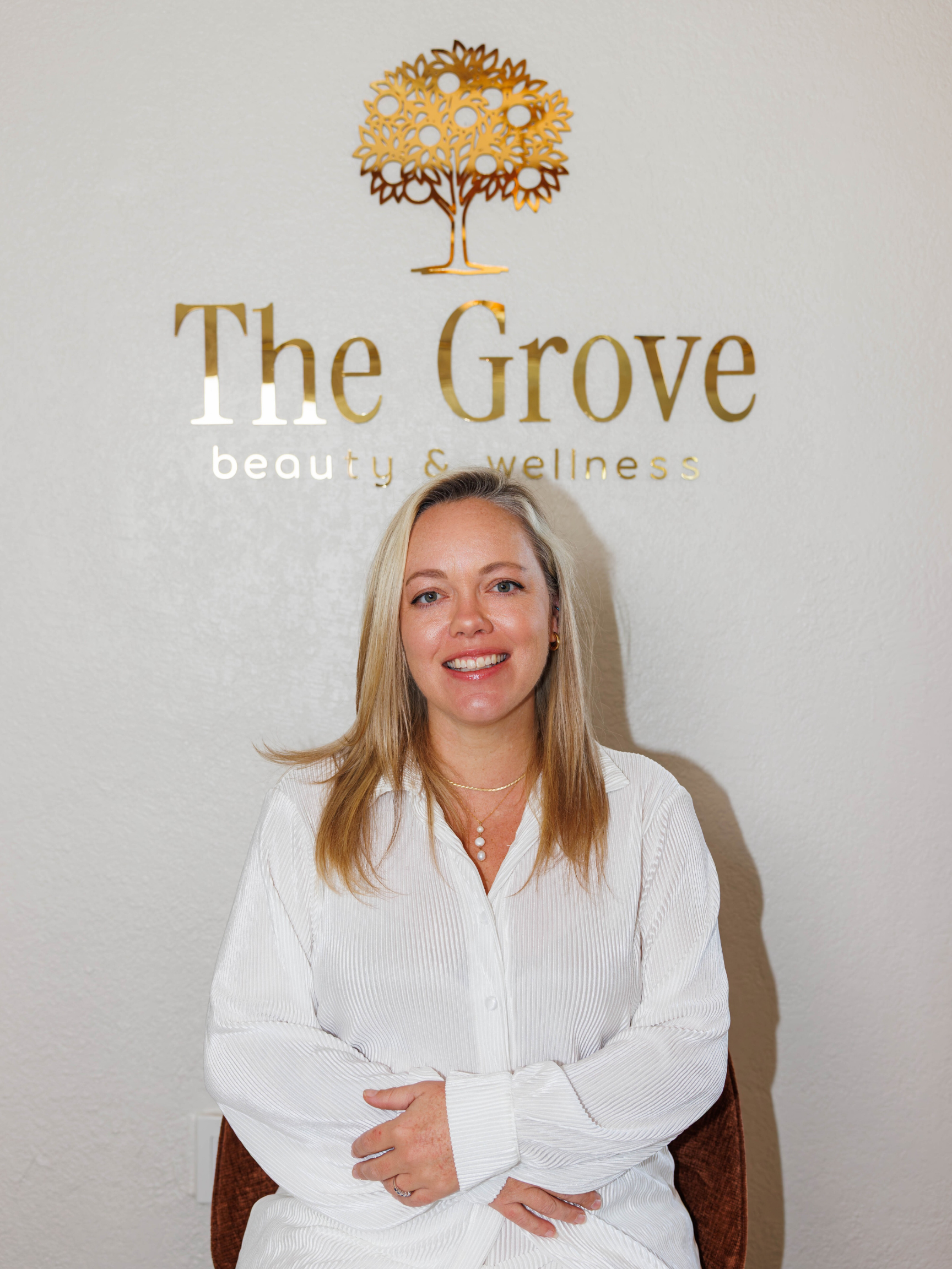 A woman sitting in front of a wall with a sign that reads 'The Grove beauty & wellness' and a decorative tree logo above the text.
