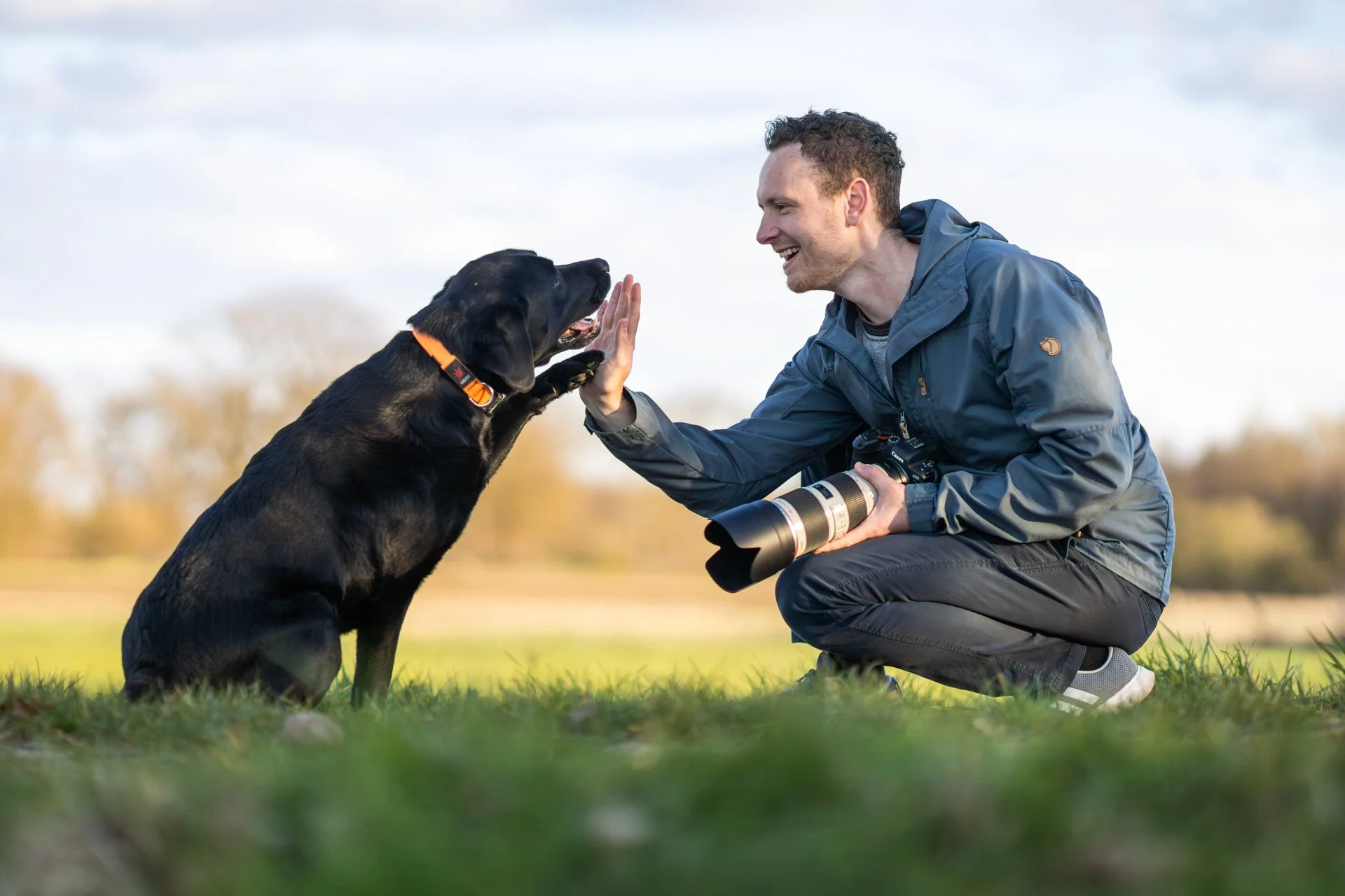 Fotograf mit Kamera in der Hand interagiert mit einem schwarzen Labrador auf einer Wiese in Hannover