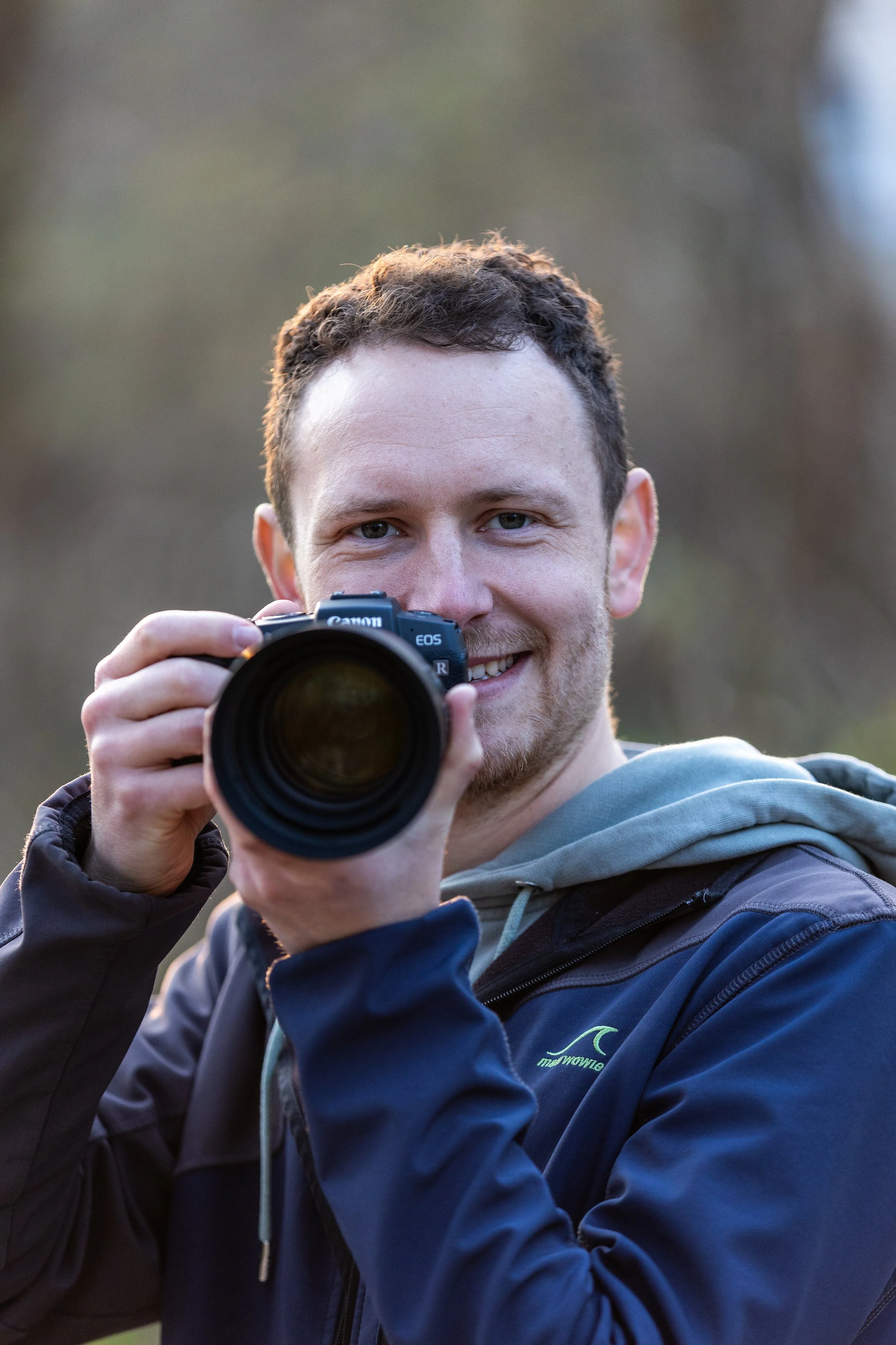 Junge Mann mit Kamera in der Hand, lächelnd, outdoor, im Hintergrund unscharfer Wald der Eilenriede in Hannover