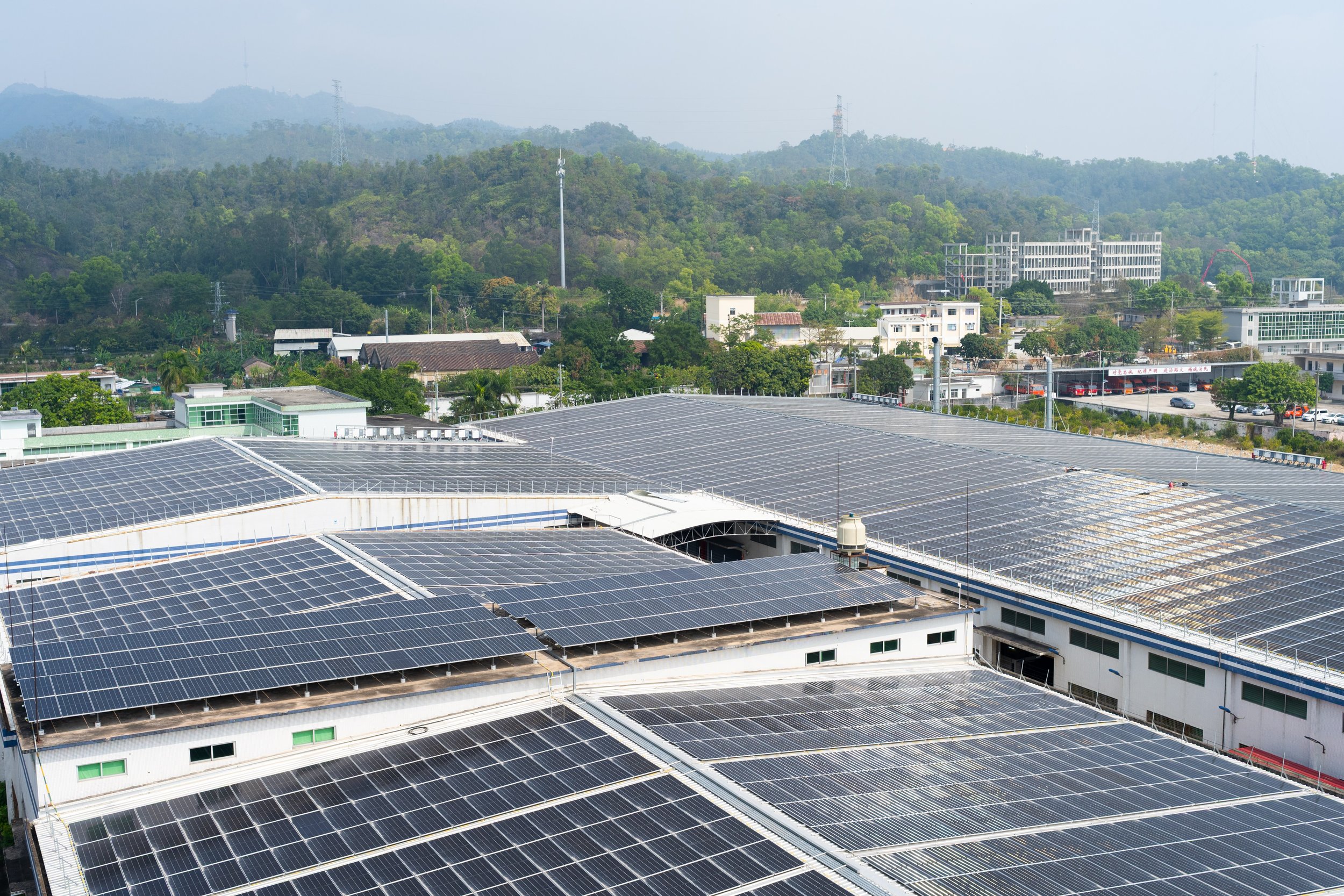 A large building covered with numerous solar panels on its roof, with a background of green hills and scattered buildings.