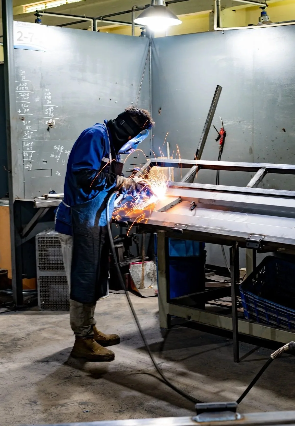 A person welding metal in a workshop with sparks flying. The worker is wearing protective gear including a face shield, gloves, and work boots, focused on their task at a metal worktable.