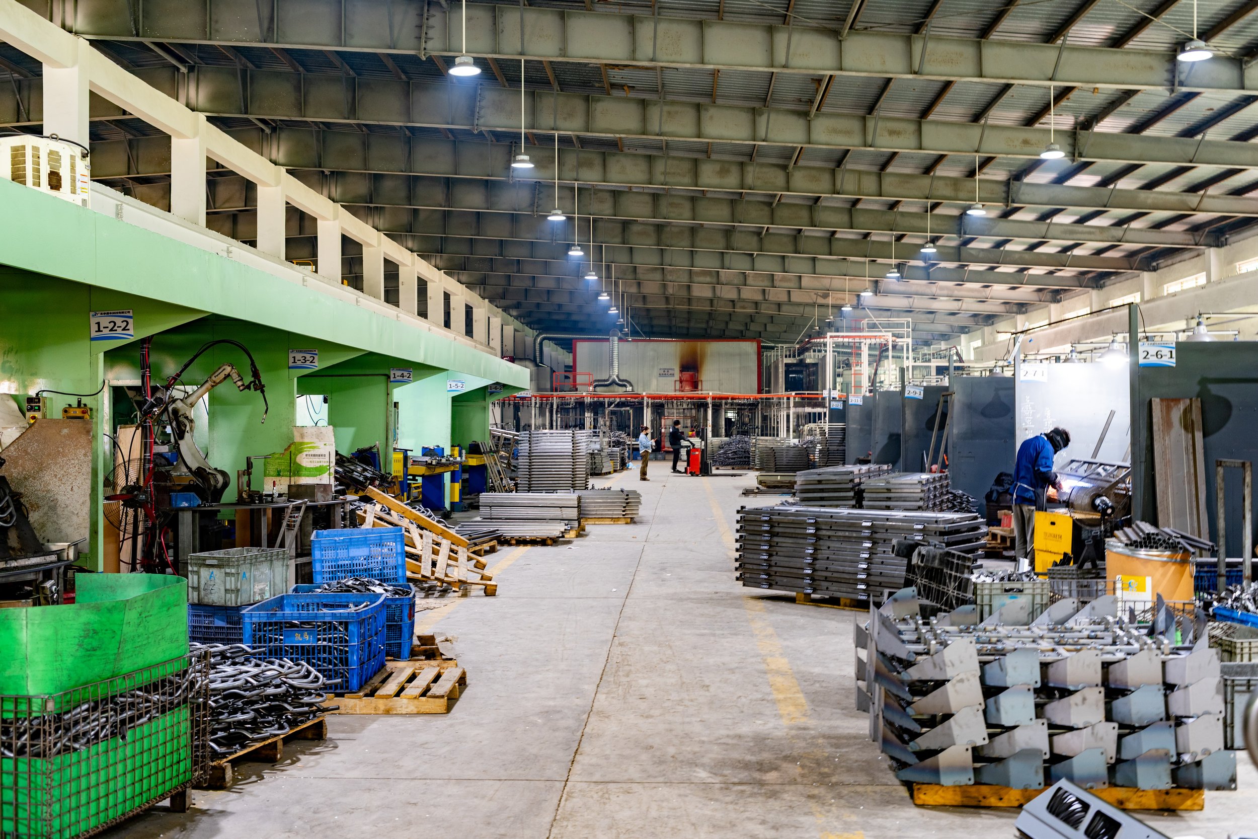 Inside an industrial manufacturing factory with machinery, workers, and metal parts organized on pallets and shelves.