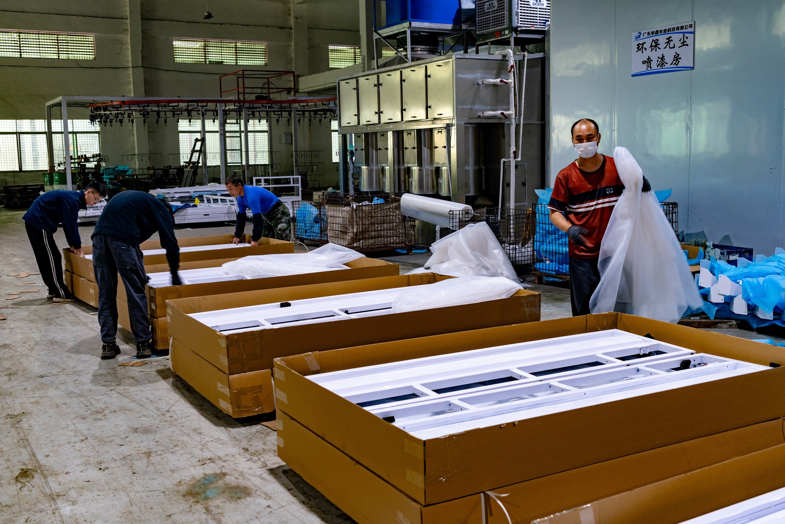 Workers assemble packaging in an industrial factory. Four workers wearing masks and gloves work around large brown boxes, preparing items for shipment.