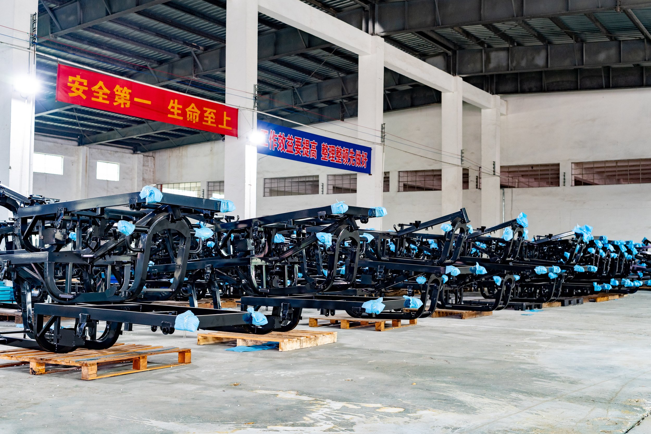 Assembly line of black metal frames for manufacturing, covered with light blue protective plastic, inside a factory with high ceilings and white walls, with red and blue Chinese safety banners hanging from the ceiling.