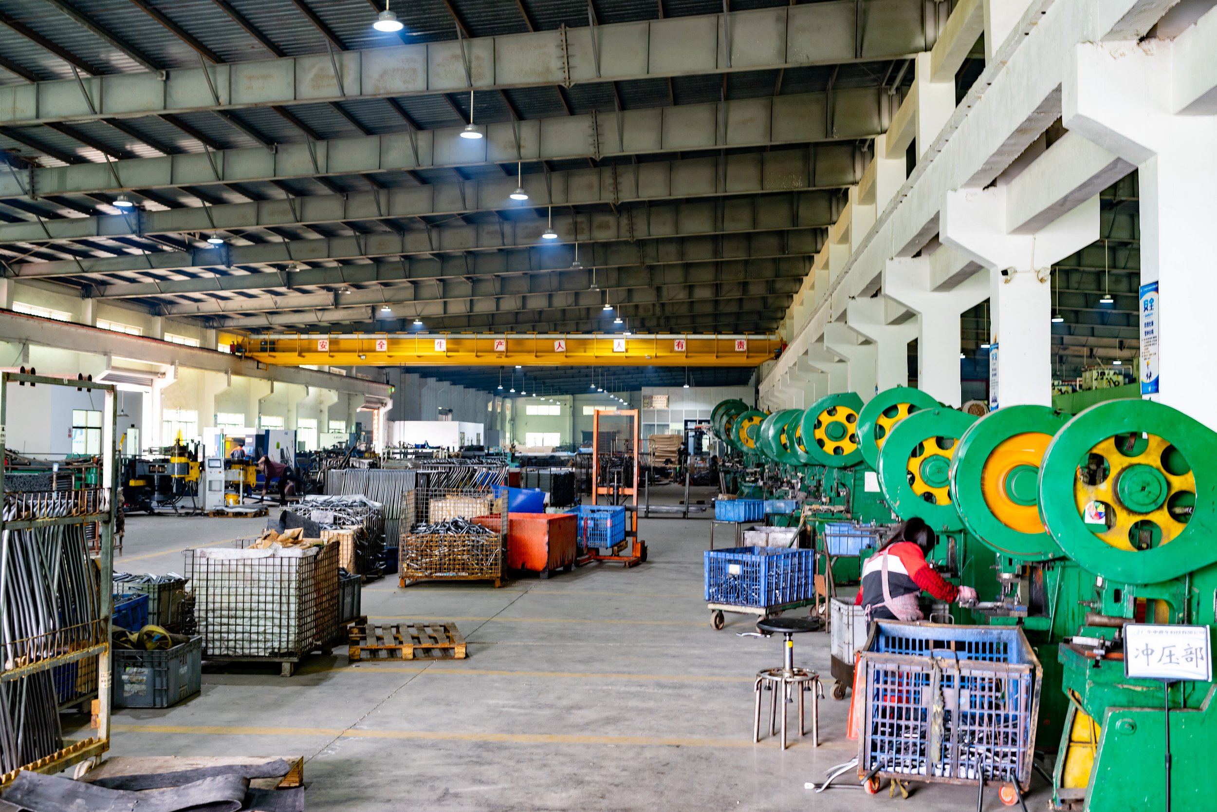 Inside a large industrial factory with multiple green and yellow machines on the right side and various carts and metal parts scattered across the gray concrete floor. Overhead lighting illuminates the spacious warehouse. A person is working near the