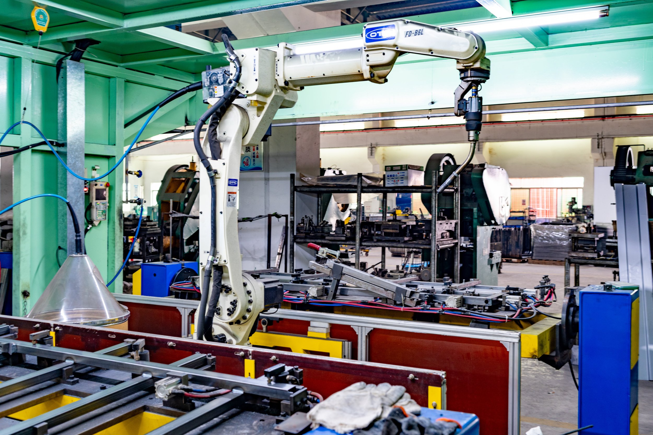 Industrial robotic arm operating in a manufacturing facility with machinery and workstations in the background.