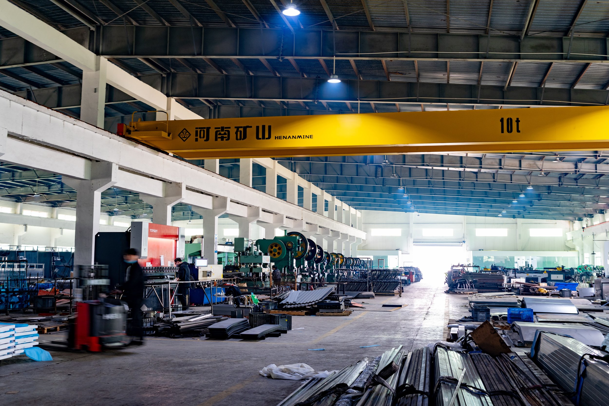 Industrial warehouse with manufacturing equipment, metal parts, and a yellow overhead crane labeled with Chinese characters and "10t" for 10 tons, under a high ceiling with bright natural light.