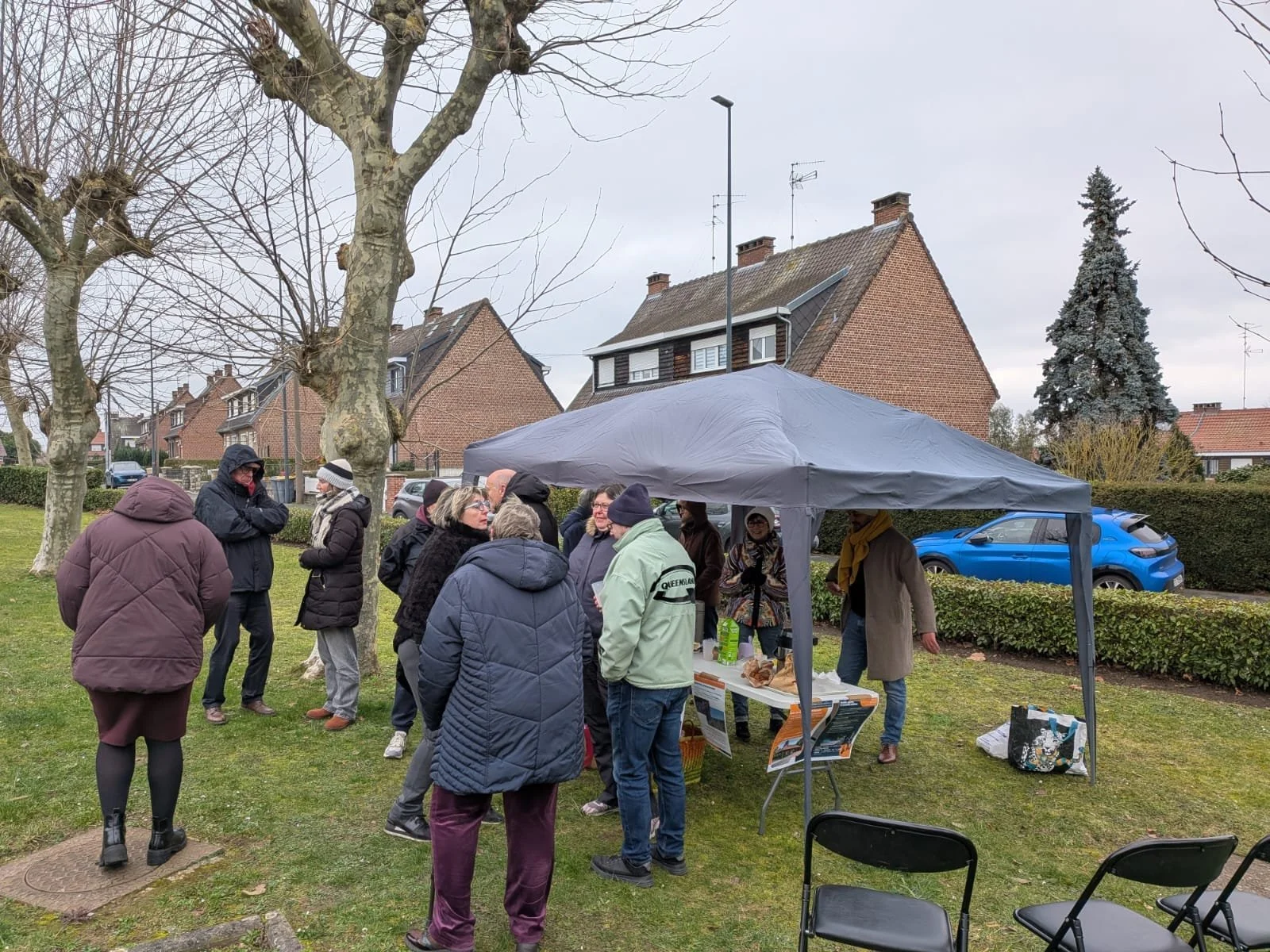 Groupe de personnes se rassemblant sous une tente dans un parc. Quelques arbres sans feuilles, maisons en arrière-plan, ciel nuageux.