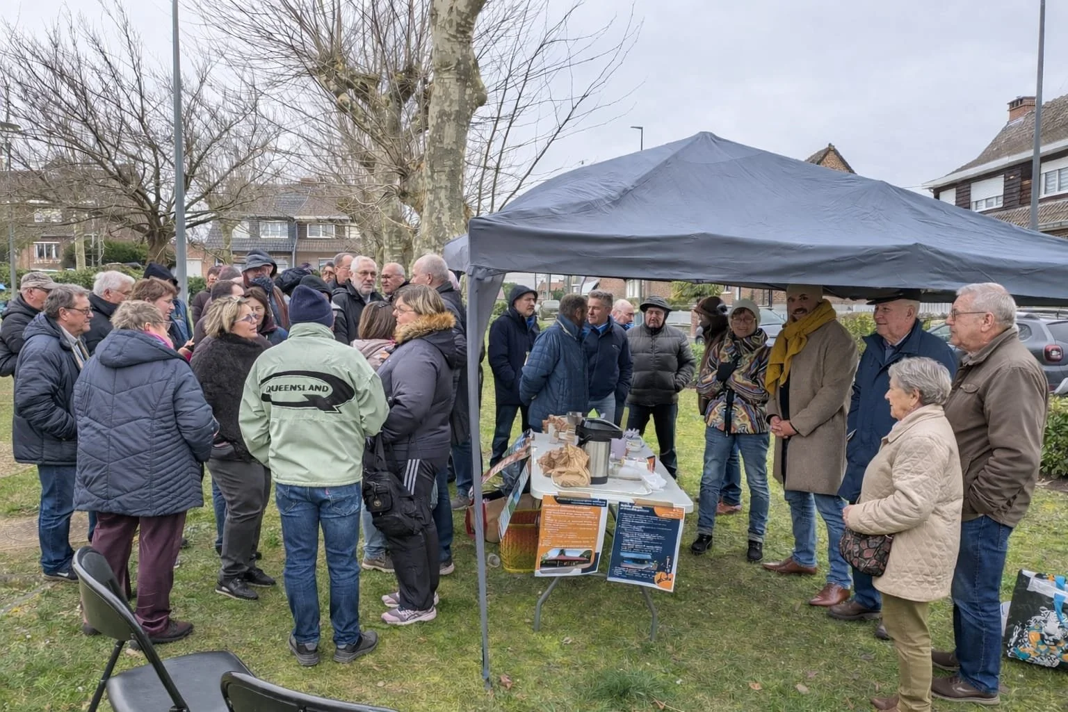 Groupe de personnes rassemblées sous une tente lors d'un événement en plein air, avec un stand proposant des documents et des boissons.