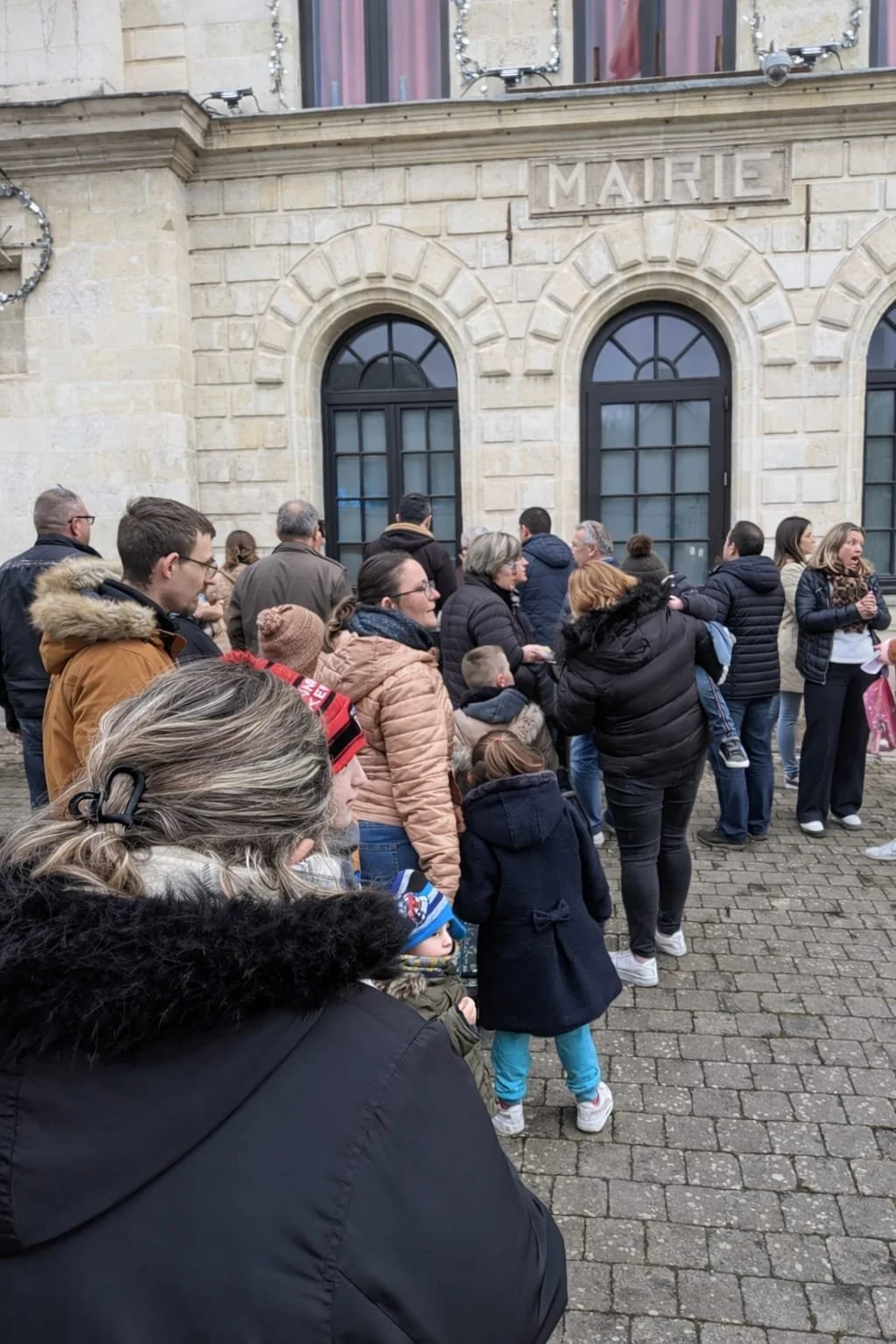 Une file d'attente de personnes devant un bâtiment en pierre avec une enseigne 'Mairie'