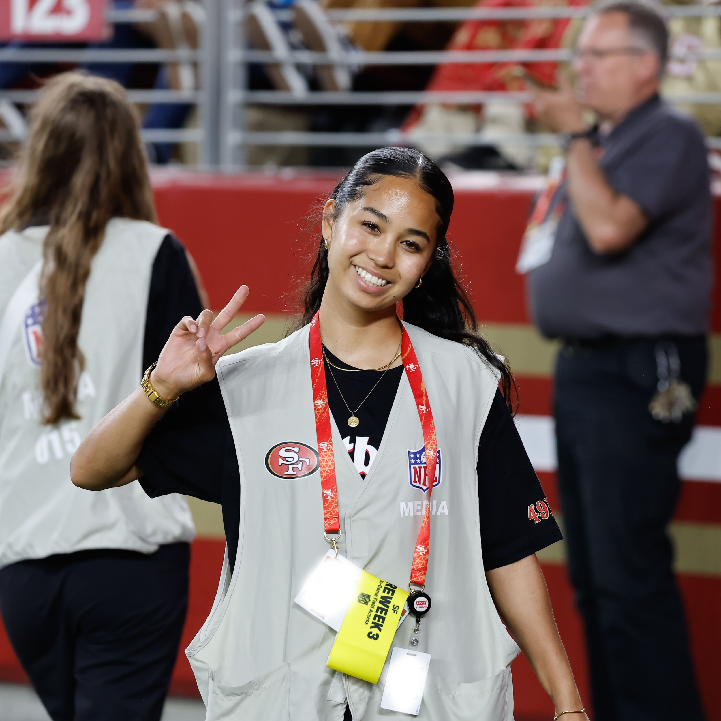 A young woman wearing a San Francisco 49ers vest and a media badge over a black shirt is smiling and making a peace sign with her right hand at a sports event. She has long, dark hair and is wearing jewelry.