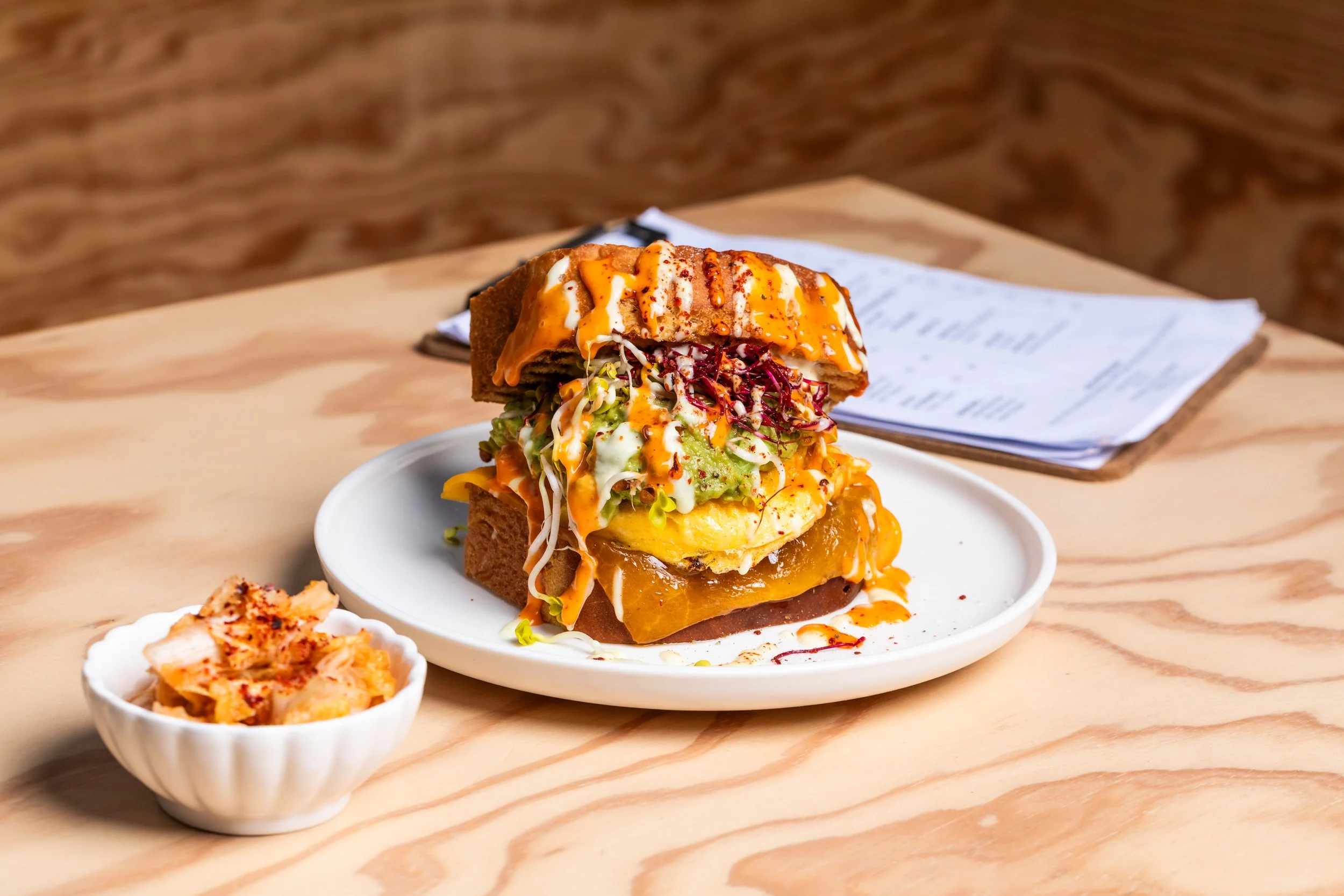 Ein mehrstöckiger Burger auf einem weißen Teller mit verschiedenen Zutaten, dazu ein Becher mit Kimchi. Der Burger enthält Salat, Avocado, eine Scheibe Käse, eine Tomate, Soßen und einen gebratenen Patties zwischen zwei Brotscheiben.
