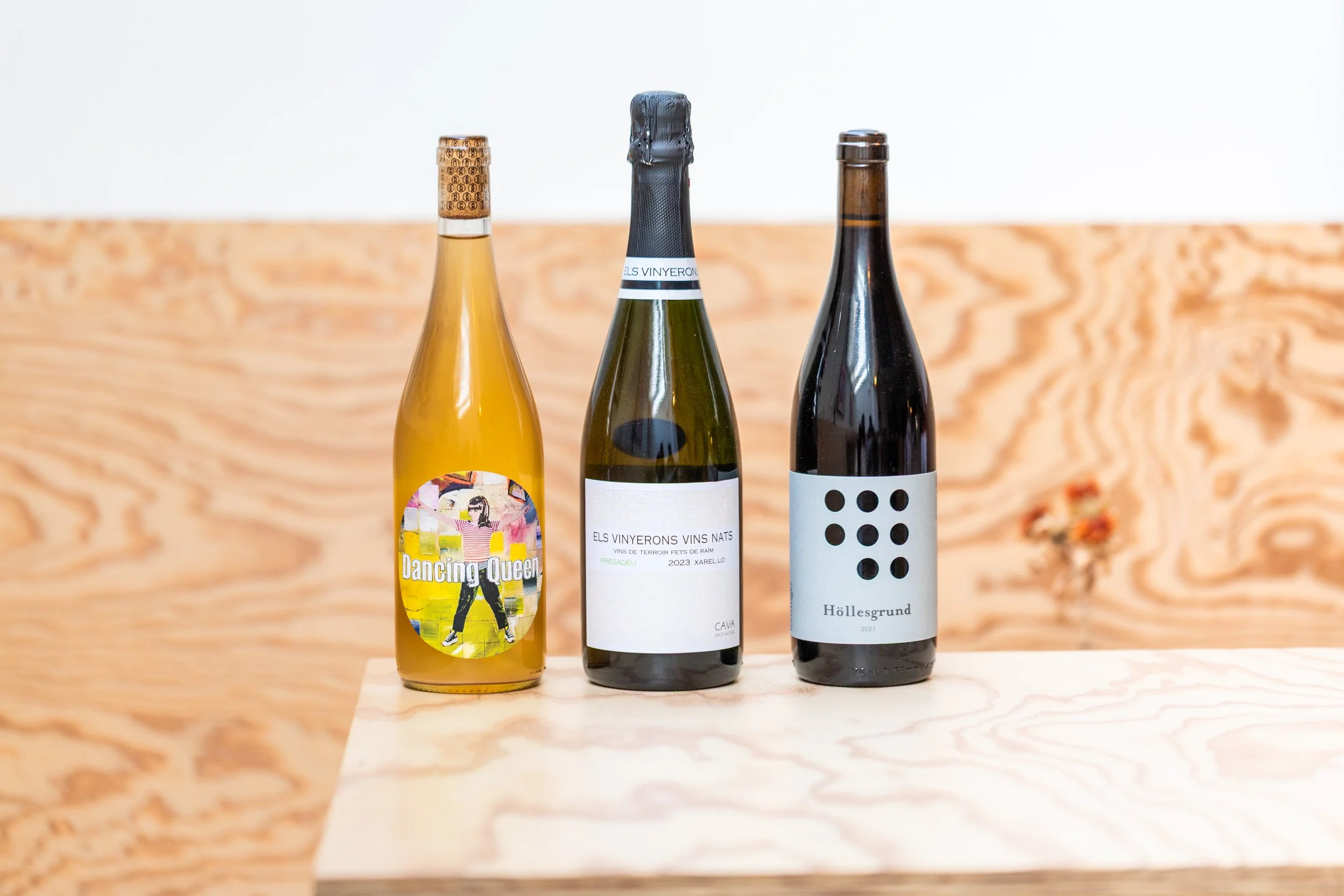 Drei Weinflaschen auf einem Holztisch, daneben eine beige Wand mit Holzmaserung.