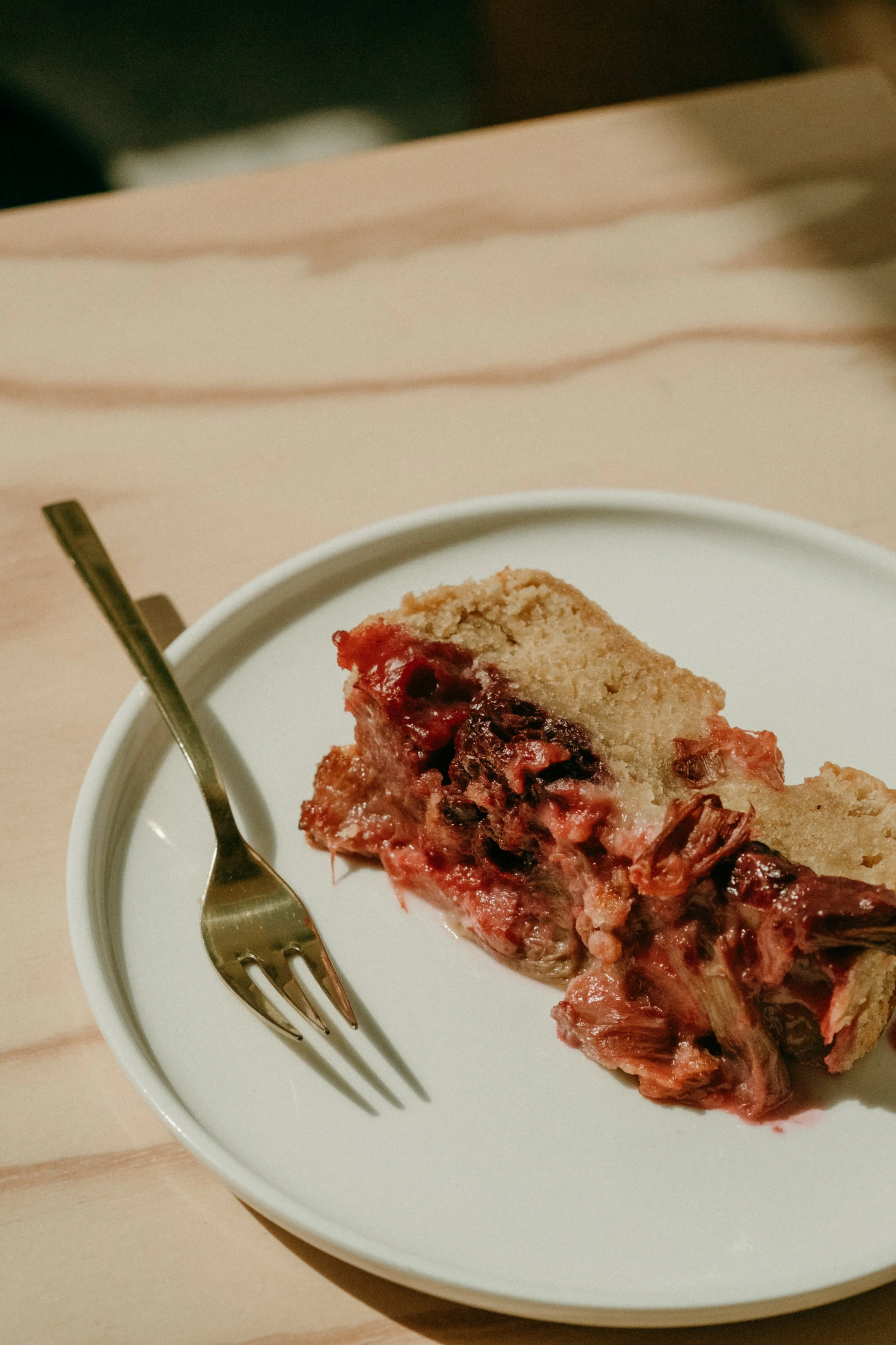 Ein Stück Kirschkuchen auf einem weißen Teller mit einer Gabel neben dem Kuchenstück, auf einem hellen Holztisch.