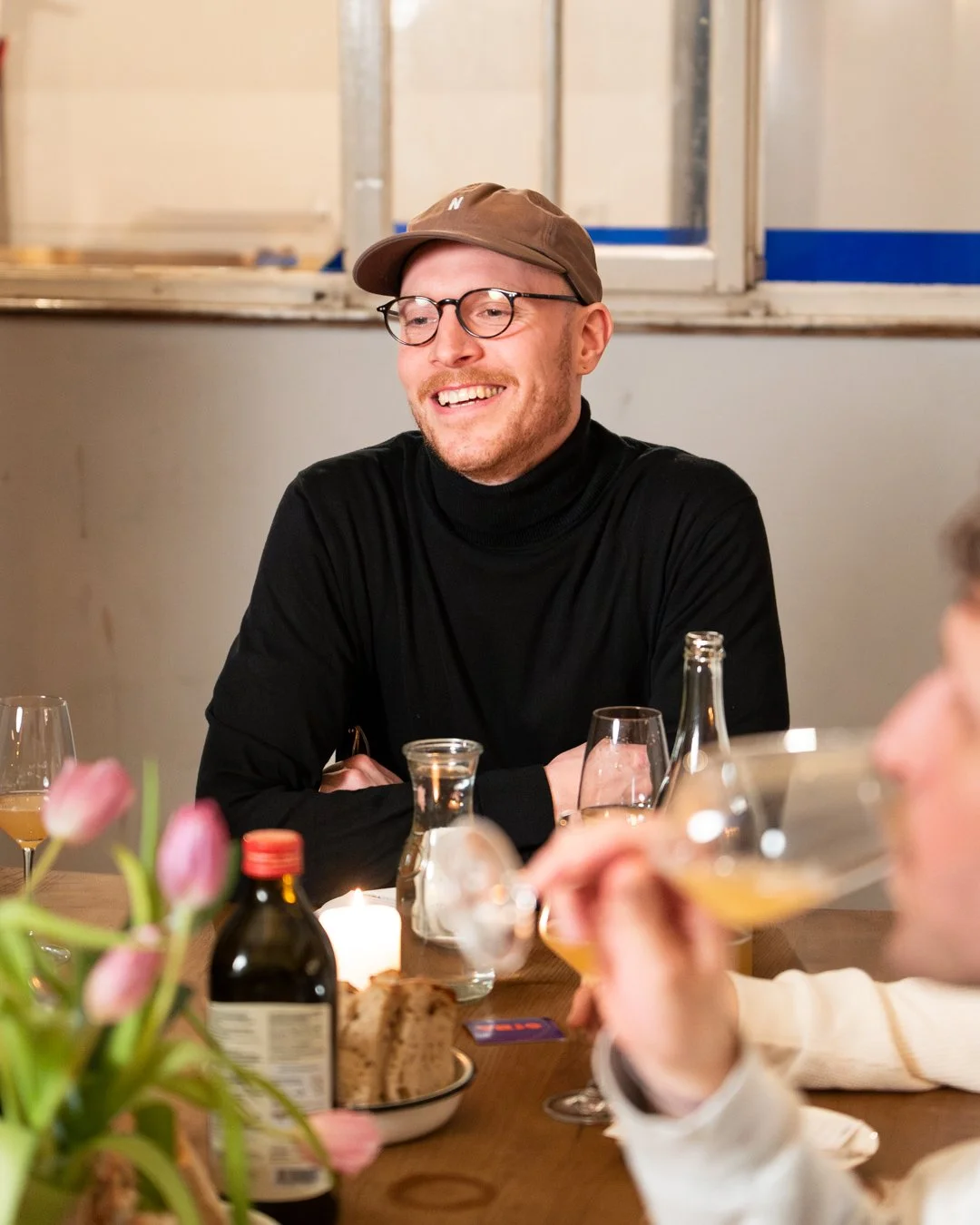Ein lachender Mann mit Brille und brauner Mütze sitzt bei einem Dinner mit mehreren Getränken, Kerzen und Blumen auf dem Tisch.