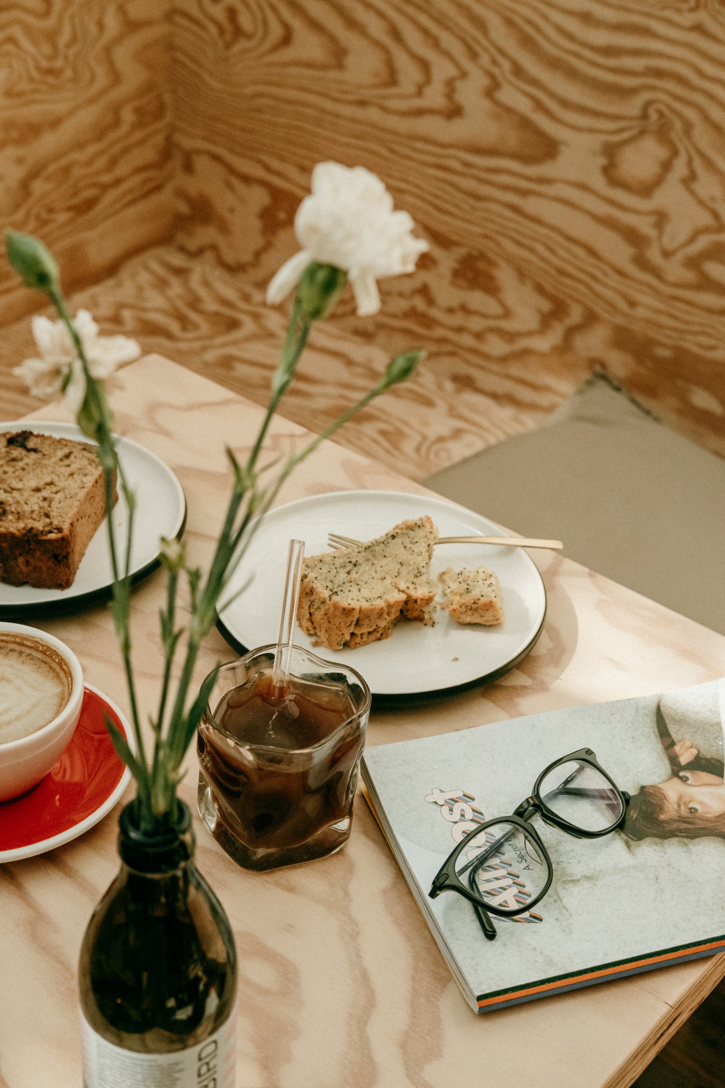 Ein Holztisch mit Teetasse, Glas mit Cola, Kuchen, Blumen und einer Brille auf einer Zeitschrift.
