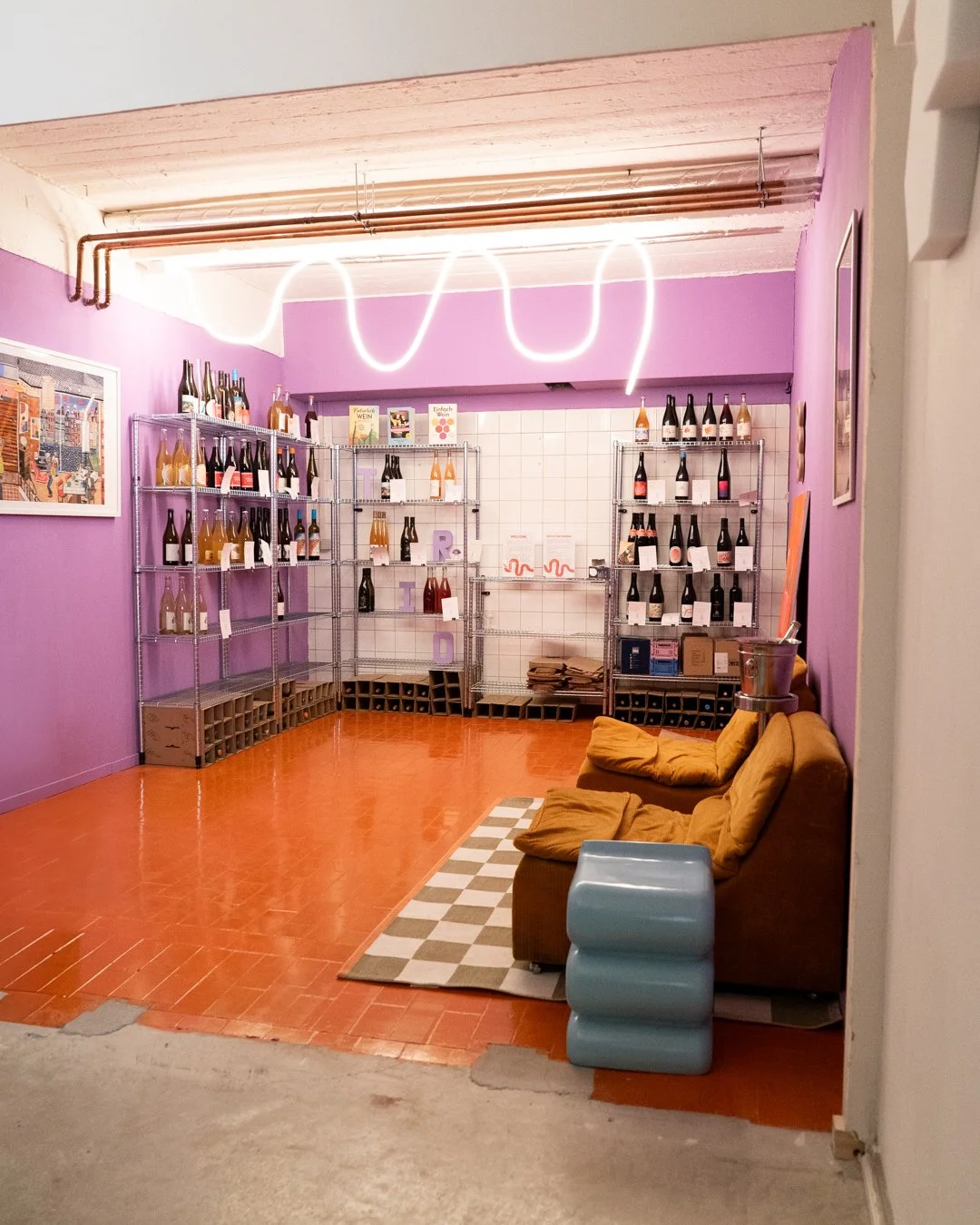 Ein Raum mit lilafarbenen Wänden, mit Weinflaschen auf Metallregalen, gelbem Sofa und einer kleinen blauen Konsole. Neonlicht an der Decke und ein gemustertes Teppich auf dem Boden.