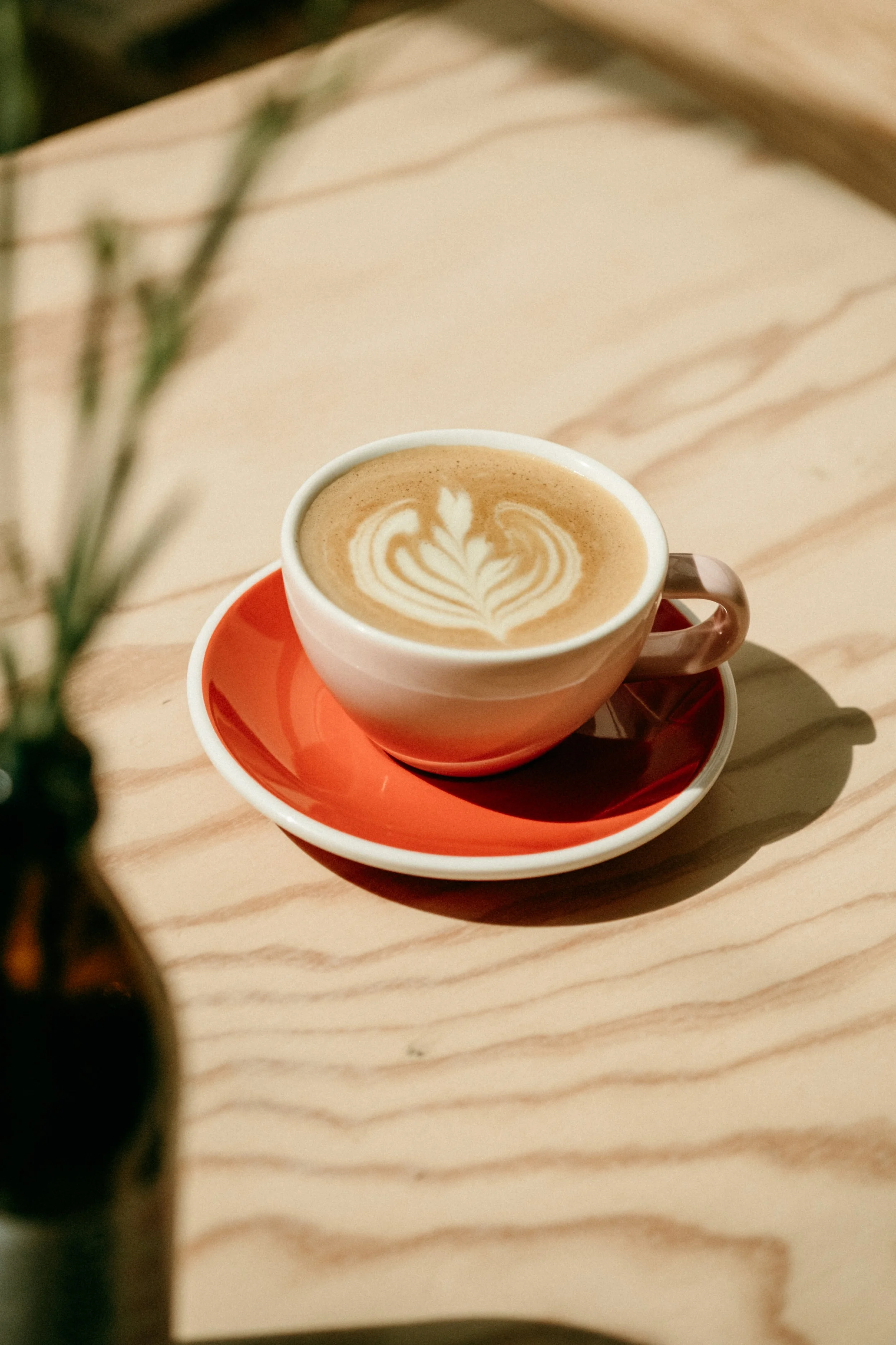 Eine Tasse Kaffee mit Latte-Art auf einem roten Unterteller, auf einem hellen Holztisch, im Vordergrund eine unscharfe Vase mit Blumen.