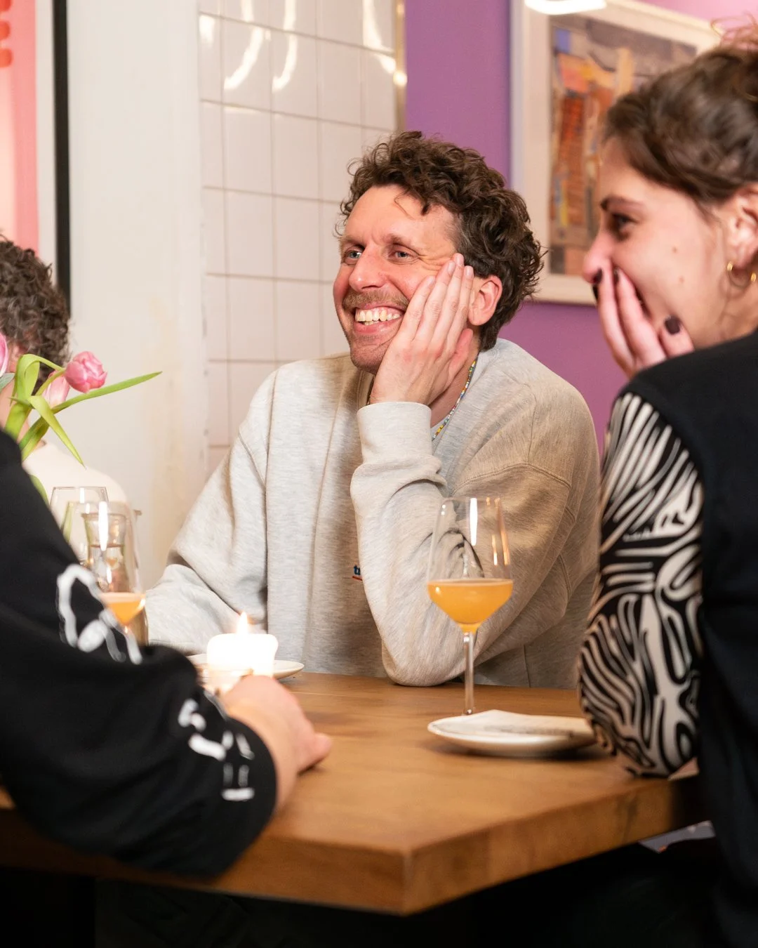 Vier Menschen sitzen an einem Tisch, lachen und unterhalten sich in einem gemütlichen Café. Es gibt ein Glas Wein, eine Kerze und eine kleine Vase mit Pinken Tulpen auf dem Tisch.