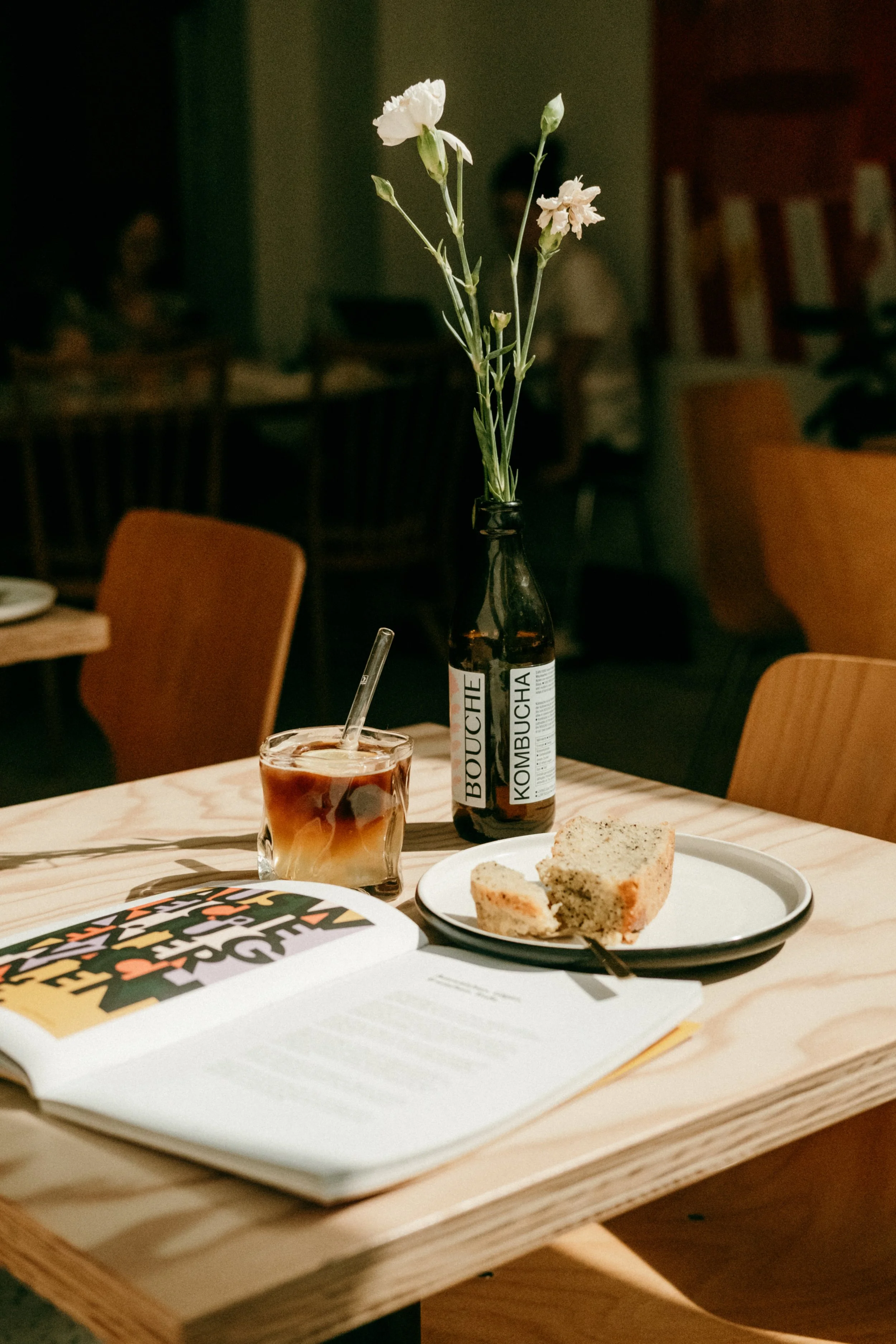 Ein Glas mit einem Kaffeegetränk, eine Vase mit weißen Blumen, ein Teller mit zwei Stück Kuchen und ein offenes Buch auf einem Holztisch in einem Restaurant.
