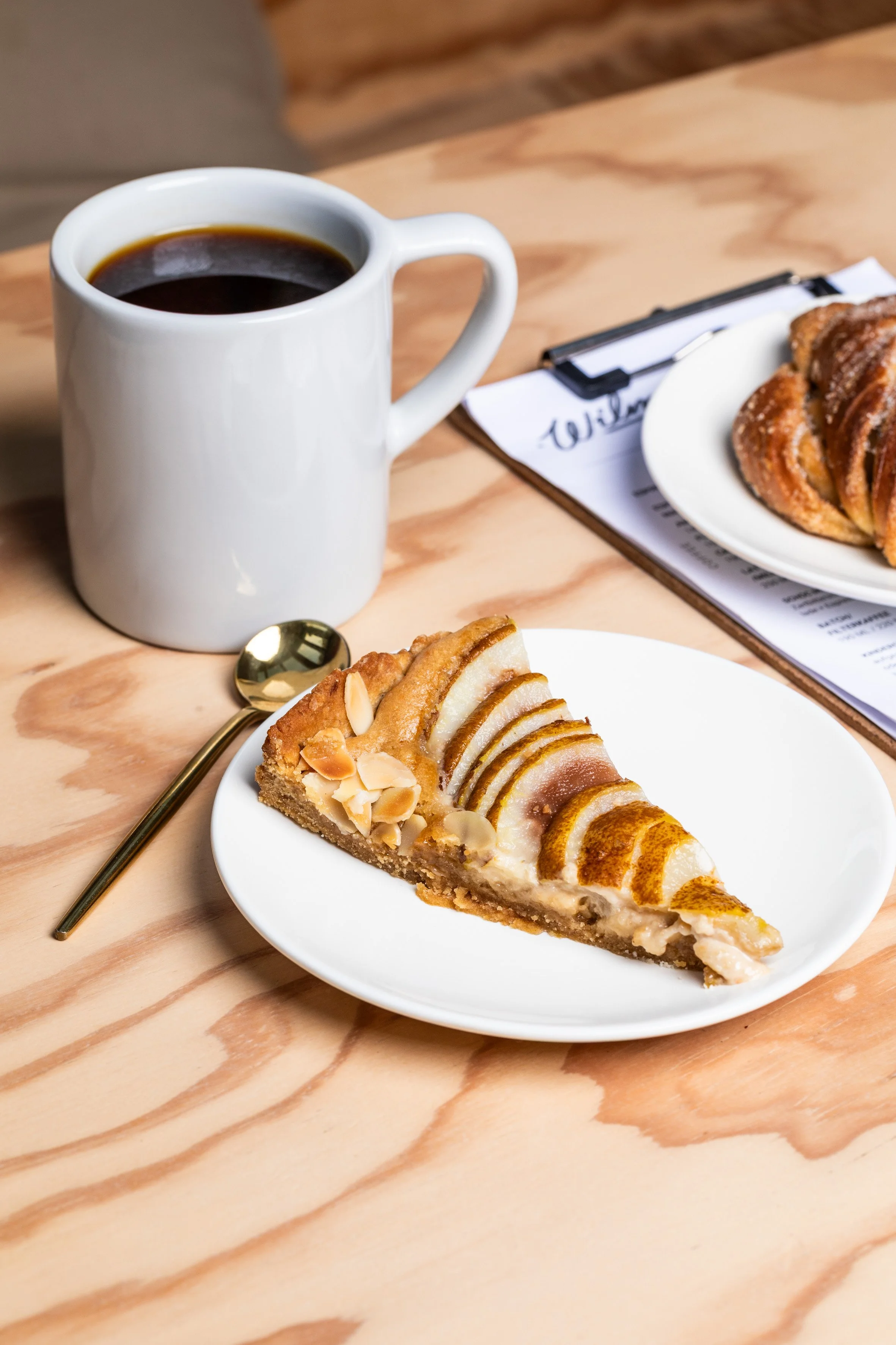 Ein Teller mit hausgebackenem Apfelkuchen, Kardamomknoten, eine Tasse mit frischem Filterkaffee, ein Löffel, ein weiterer Teller mit Gebäck und ein Klemmbrett mit Papier auf einem Holztisch.