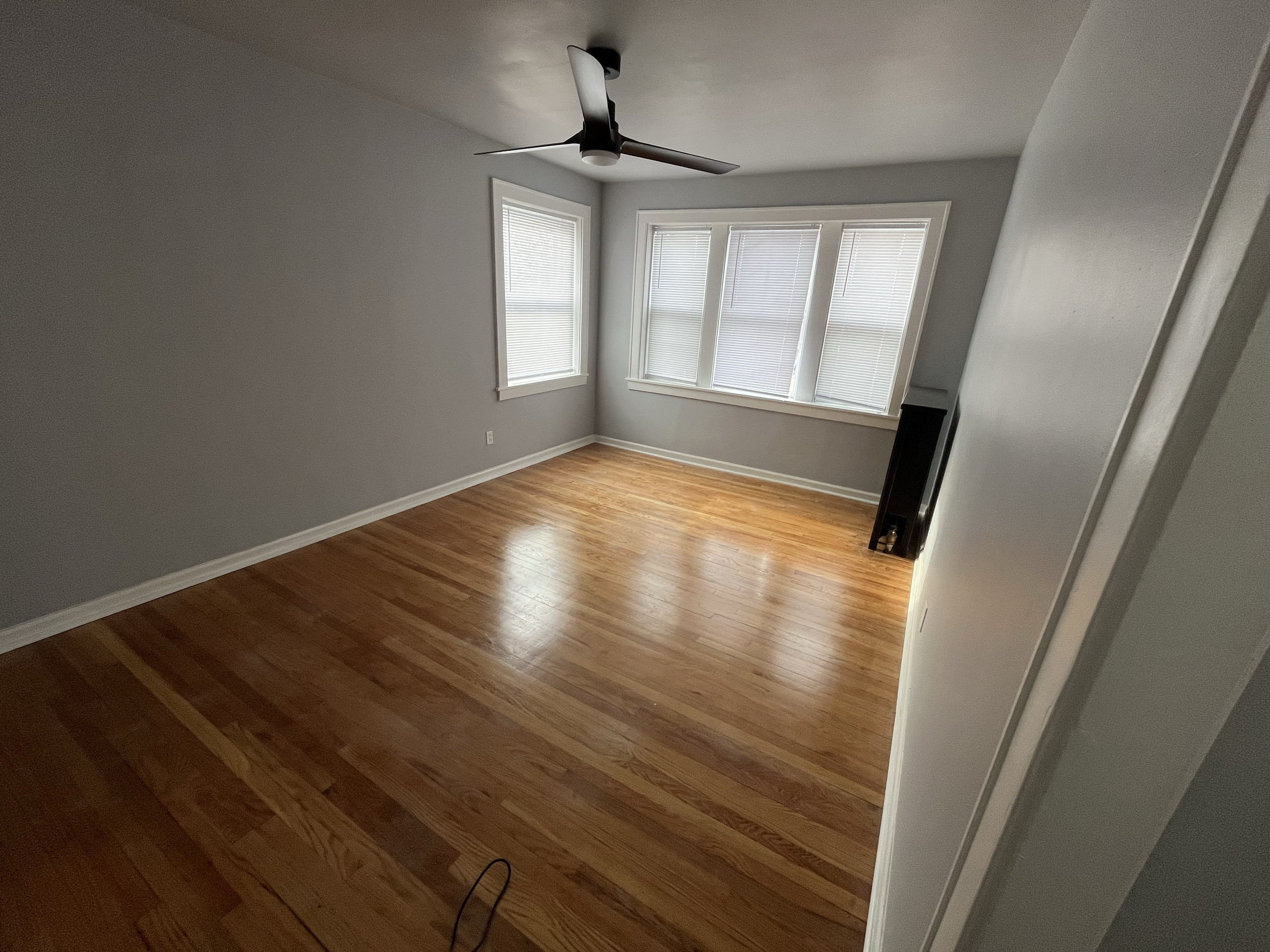 Empty room with hardwood floors, white walls, a ceiling fan, and three large windows with blinds.