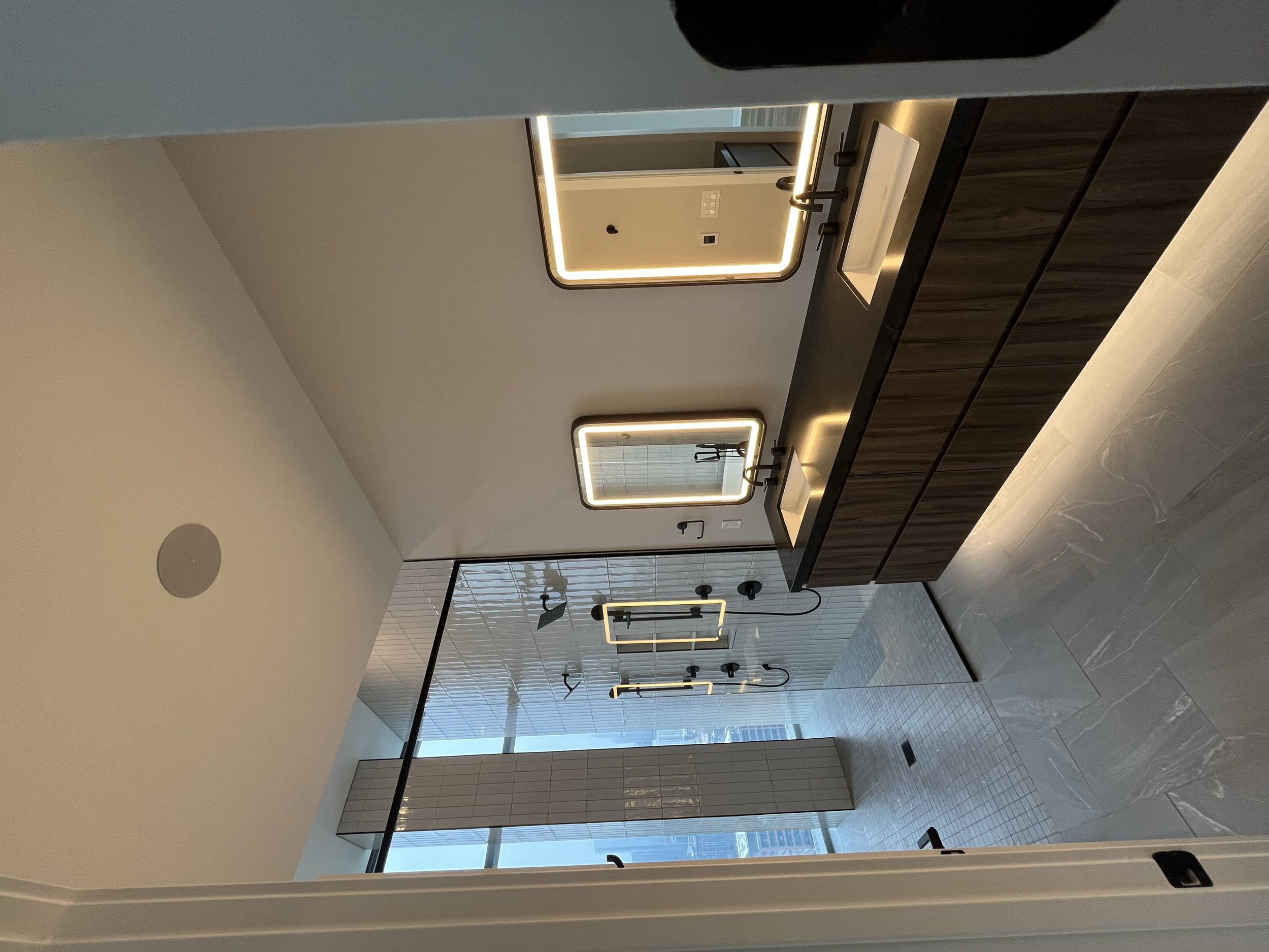 Modern bathroom with double vanity, illuminated mirrors, black countertop, and large window with city view.