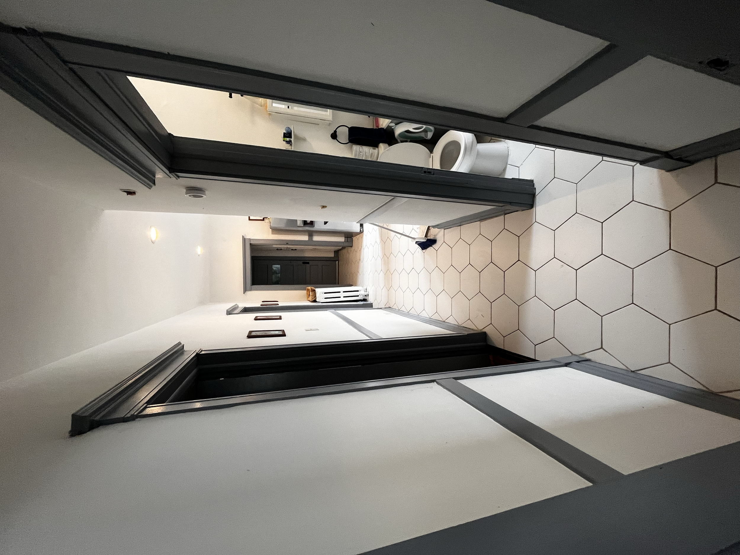 View of a hallway leading to a bathroom with open door. Visible bathroom toilet and some toiletries. Tiled floor and white walls.