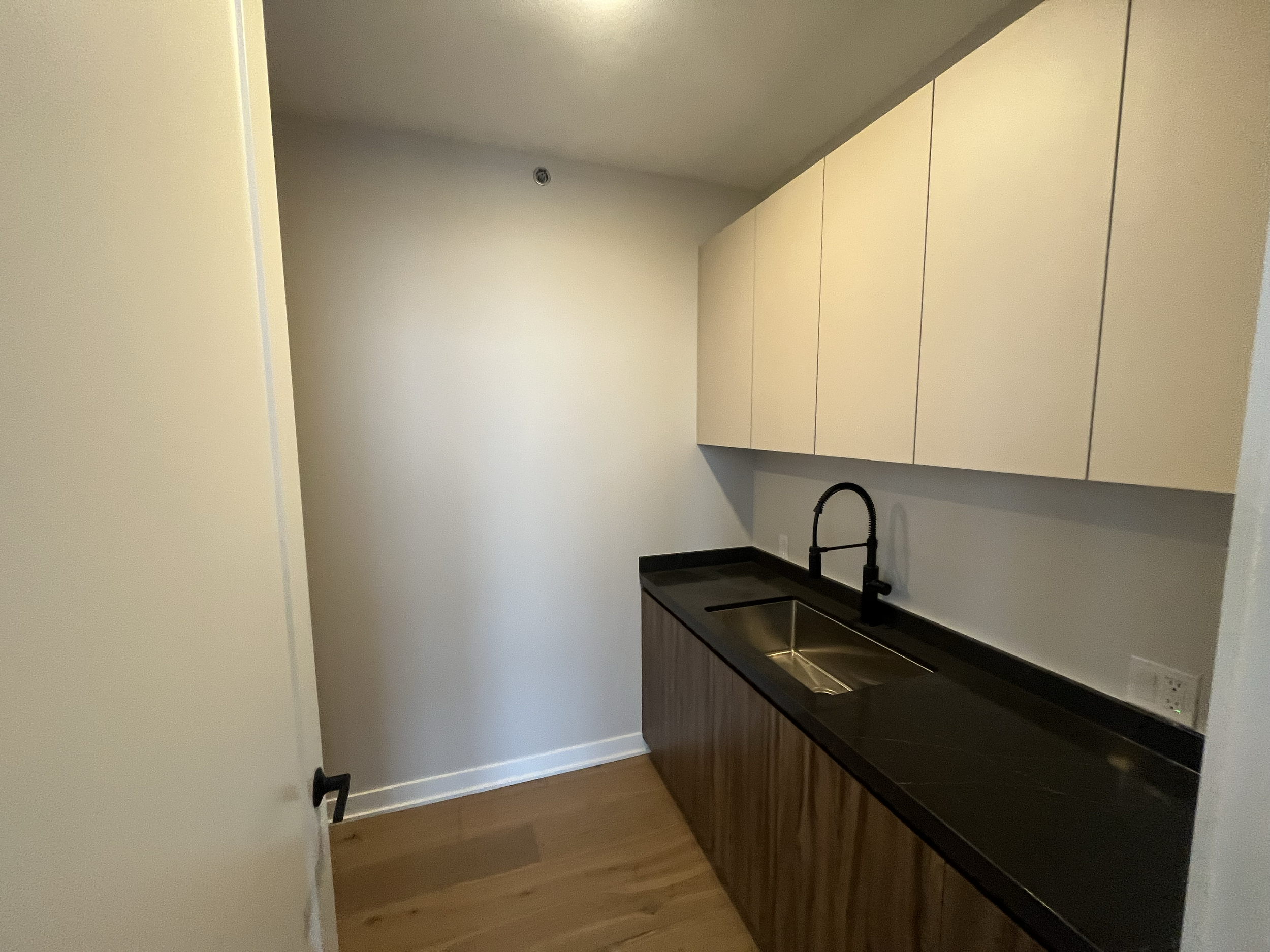 Image of a modern, minimalist kitchen with black countertop, stainless steel sink, white upper cabinets, and wooden lower cabinets, with hardwood flooring.
