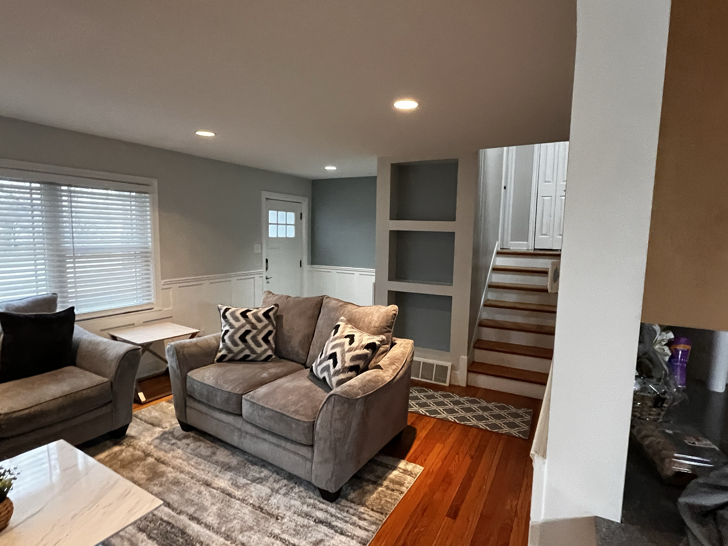 Living room with gray sofas, patterned pillows, window blinds, hardwood floors, stairs, and built-in shelves.