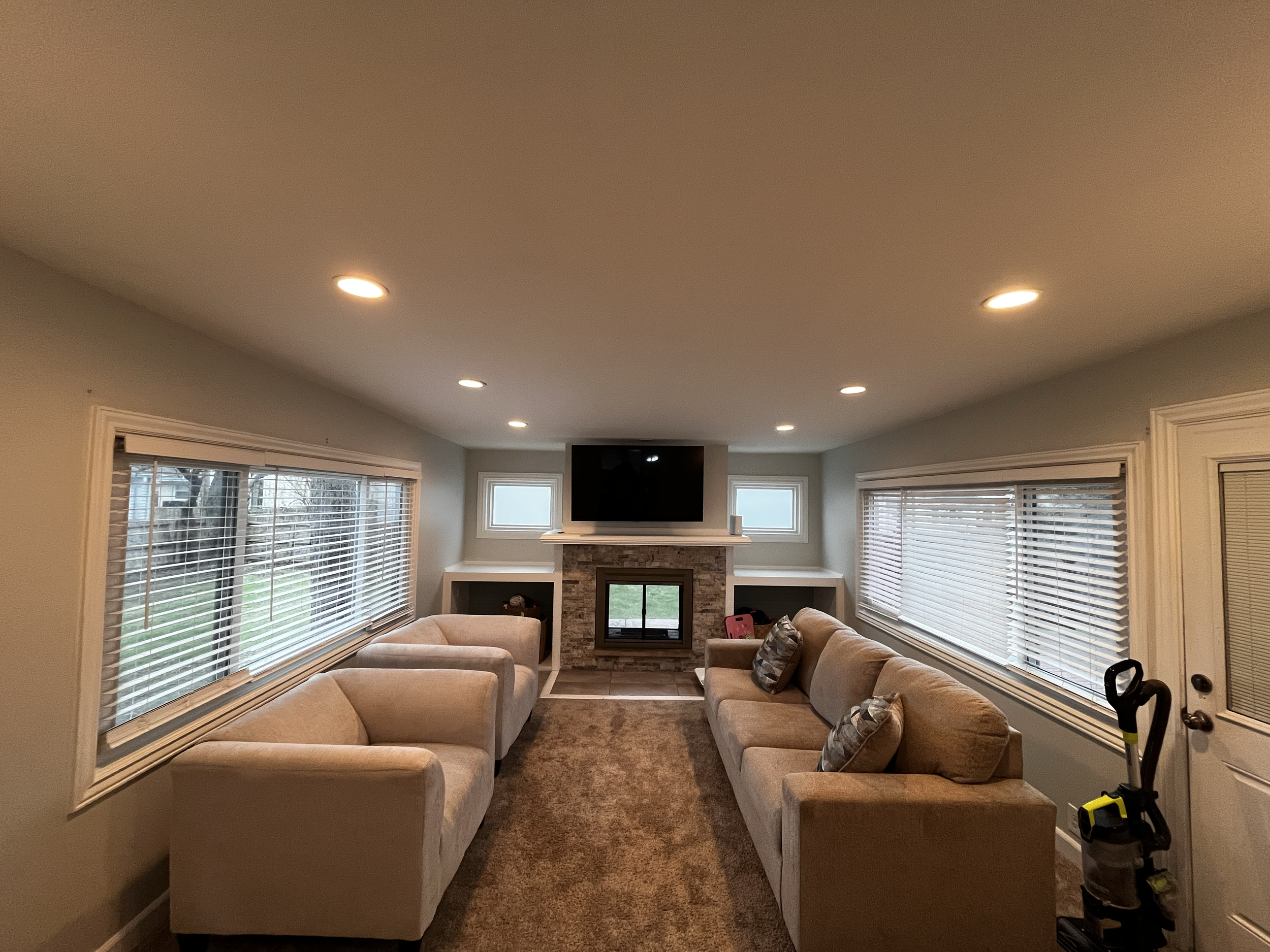 Living room with beige sofas, a fireplace with a mounted TV above it, large windows with blinds, and a carpeted floor.