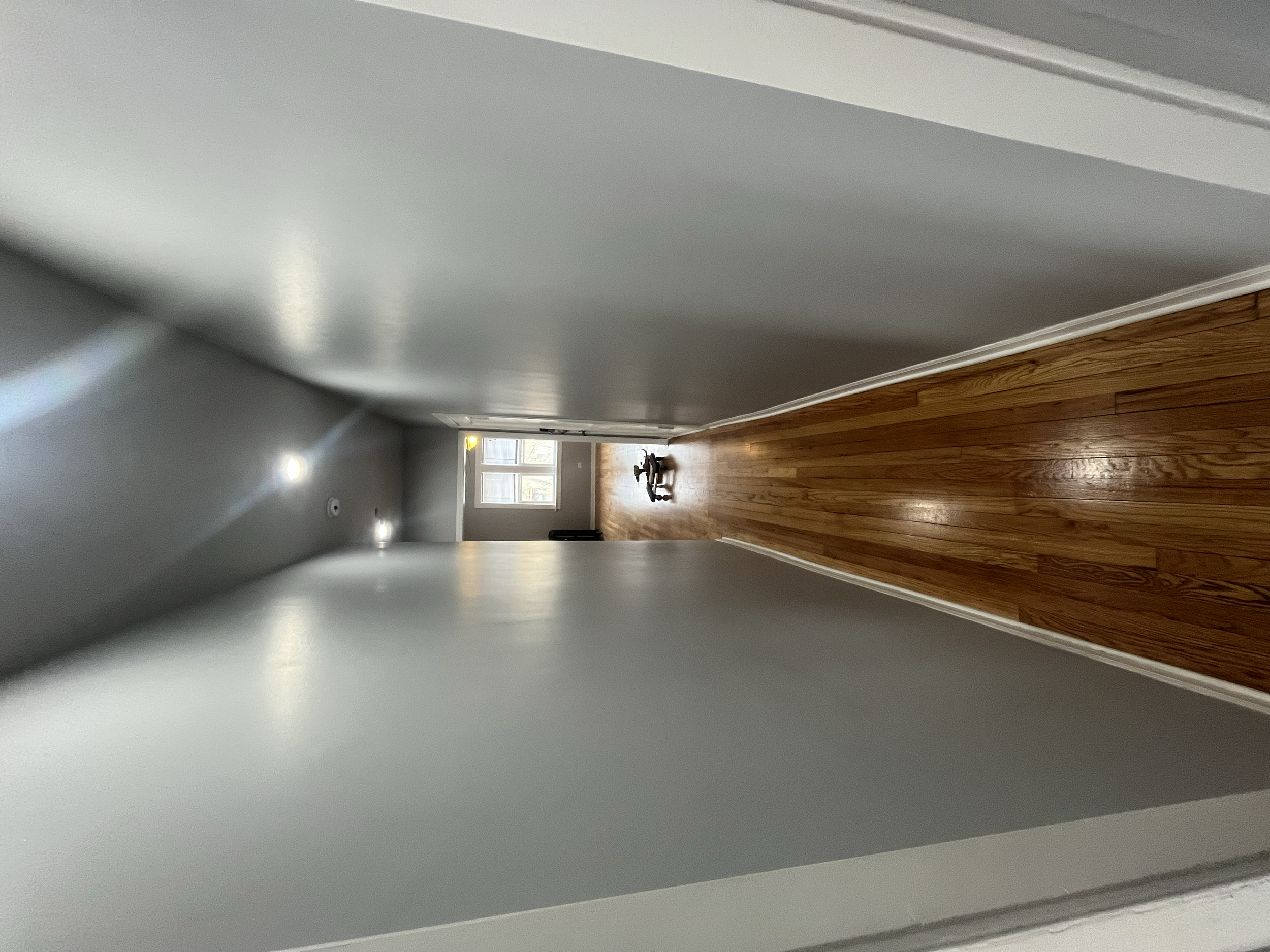 A view down a hallway with gray walls, hardwood floors, a window at the end, and a small piece of artwork on the floor near the window.