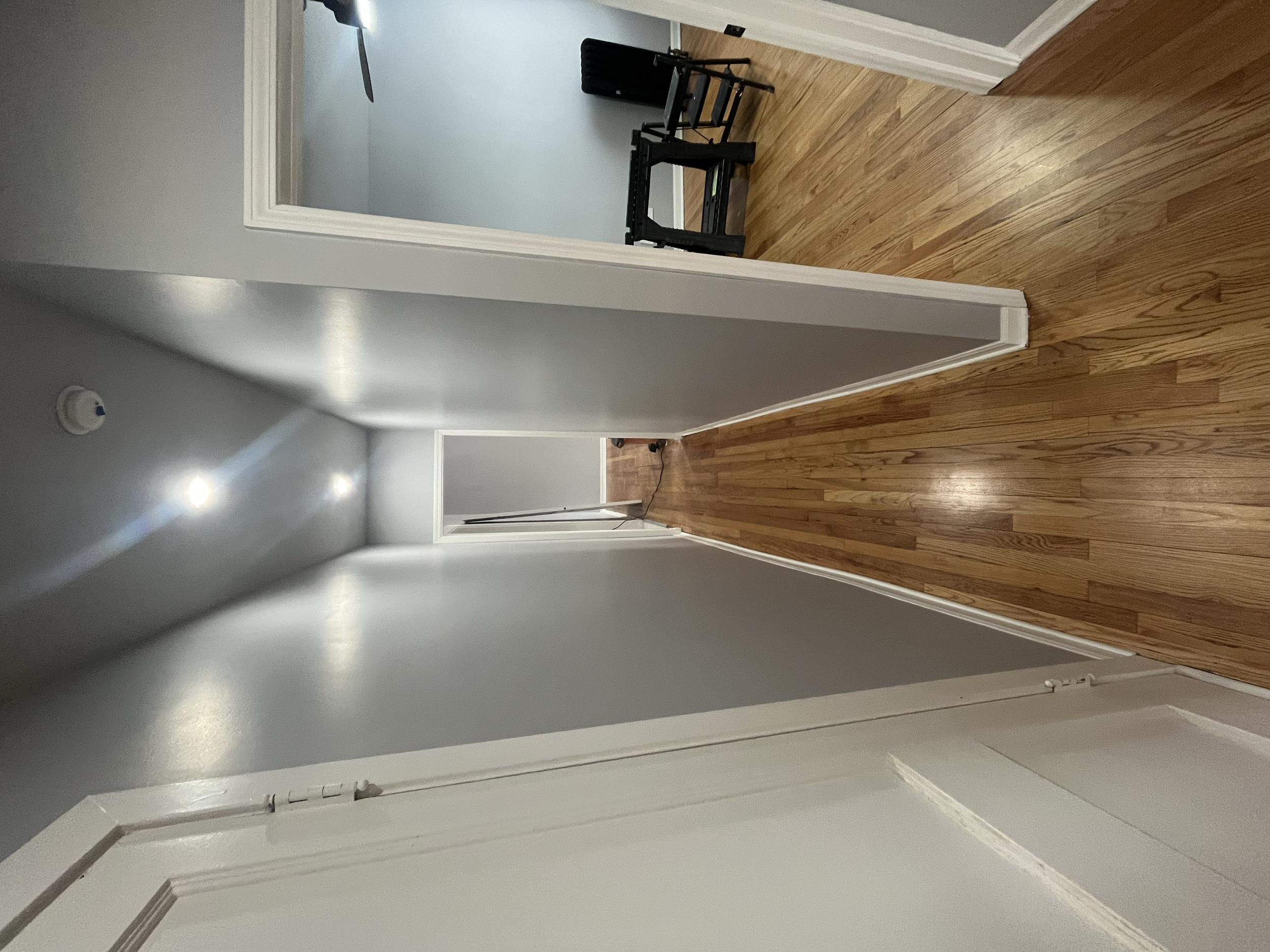 Interior hallway with hardwood flooring, white walls, and a doorway leading to a room with light blue walls and a black bench.