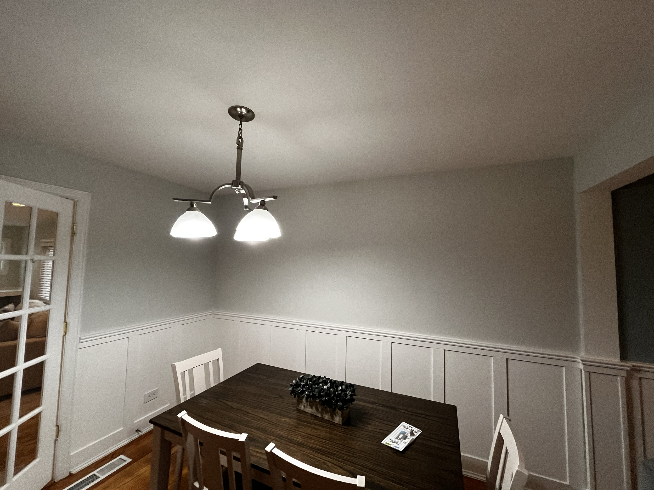 Dining room with a dark wood table, white chairs, a decorative plant, and a modern ceiling light fixture with two lamps