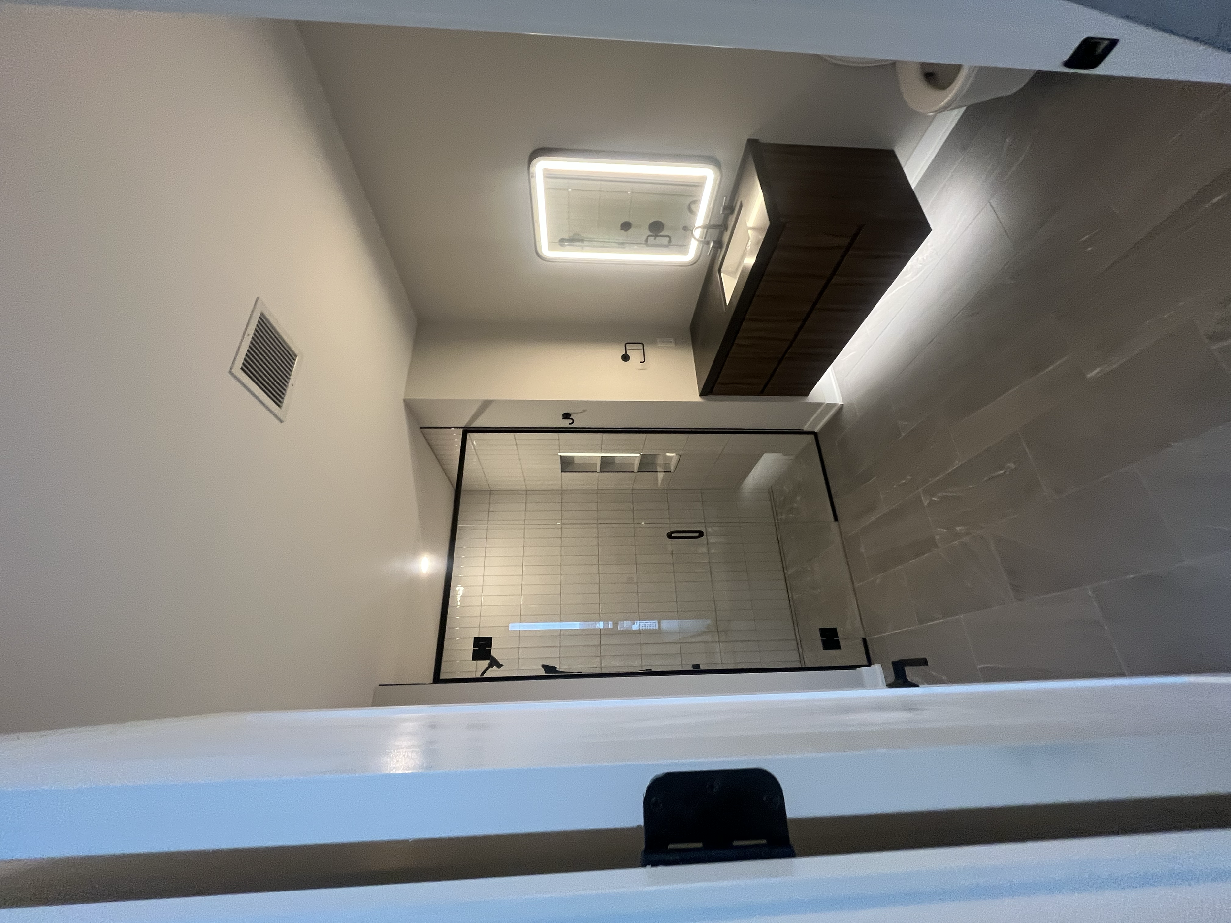 Modern bathroom with tiled shower, small wooden cabinet, and illuminated mirror.