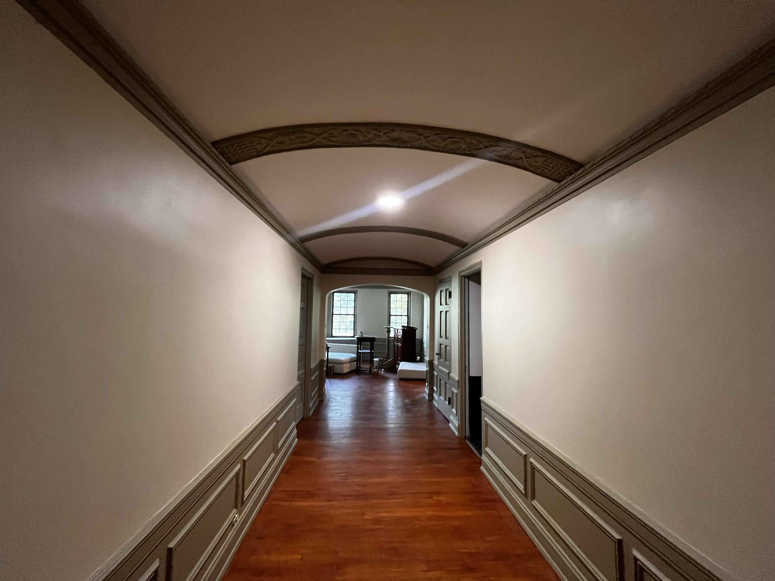 A hallway with wooden floors, cream-colored walls with decorative wainscoting, and an arched ceiling with wooden beams. At the end of the hallway, there is a room with large windows and furniture.