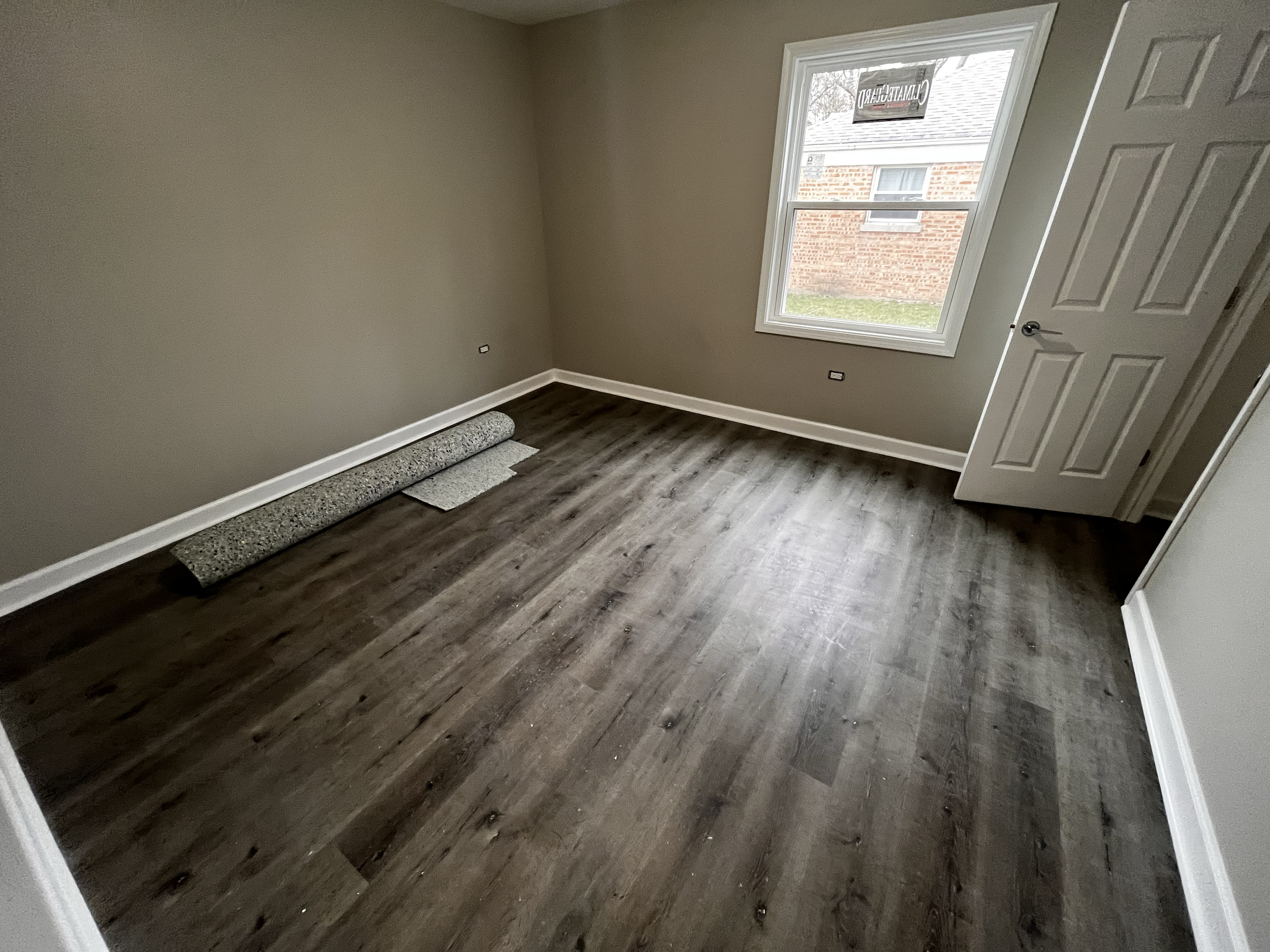 Empty room with beige walls, a window, and brown wood flooring. There is a mat and a rolled-up carpet near the wall, and an open door showing part of a closet.