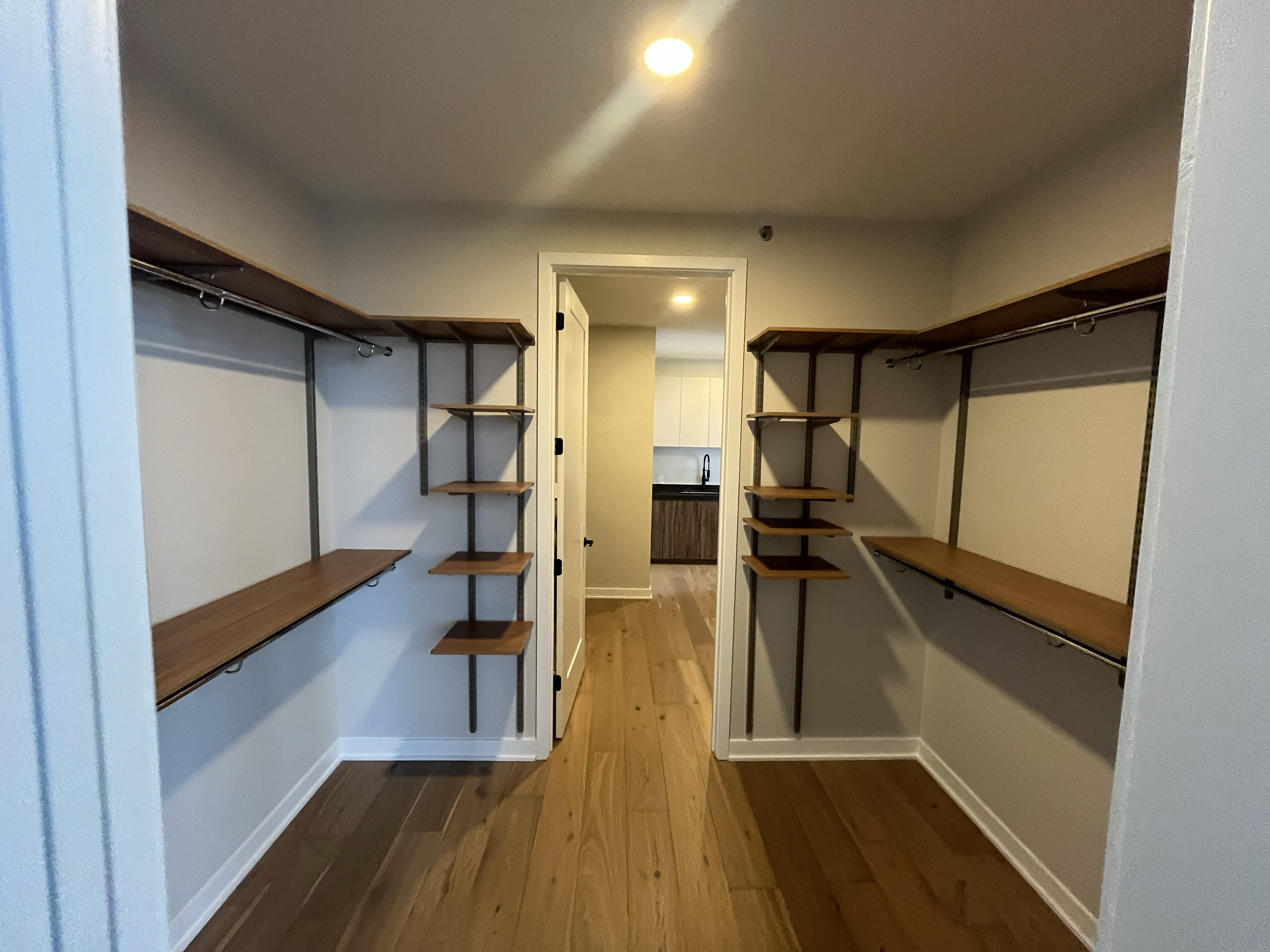 Empty walk-in closet with wooden shelves on both sides and a view into a small kitchen area through an open door.
