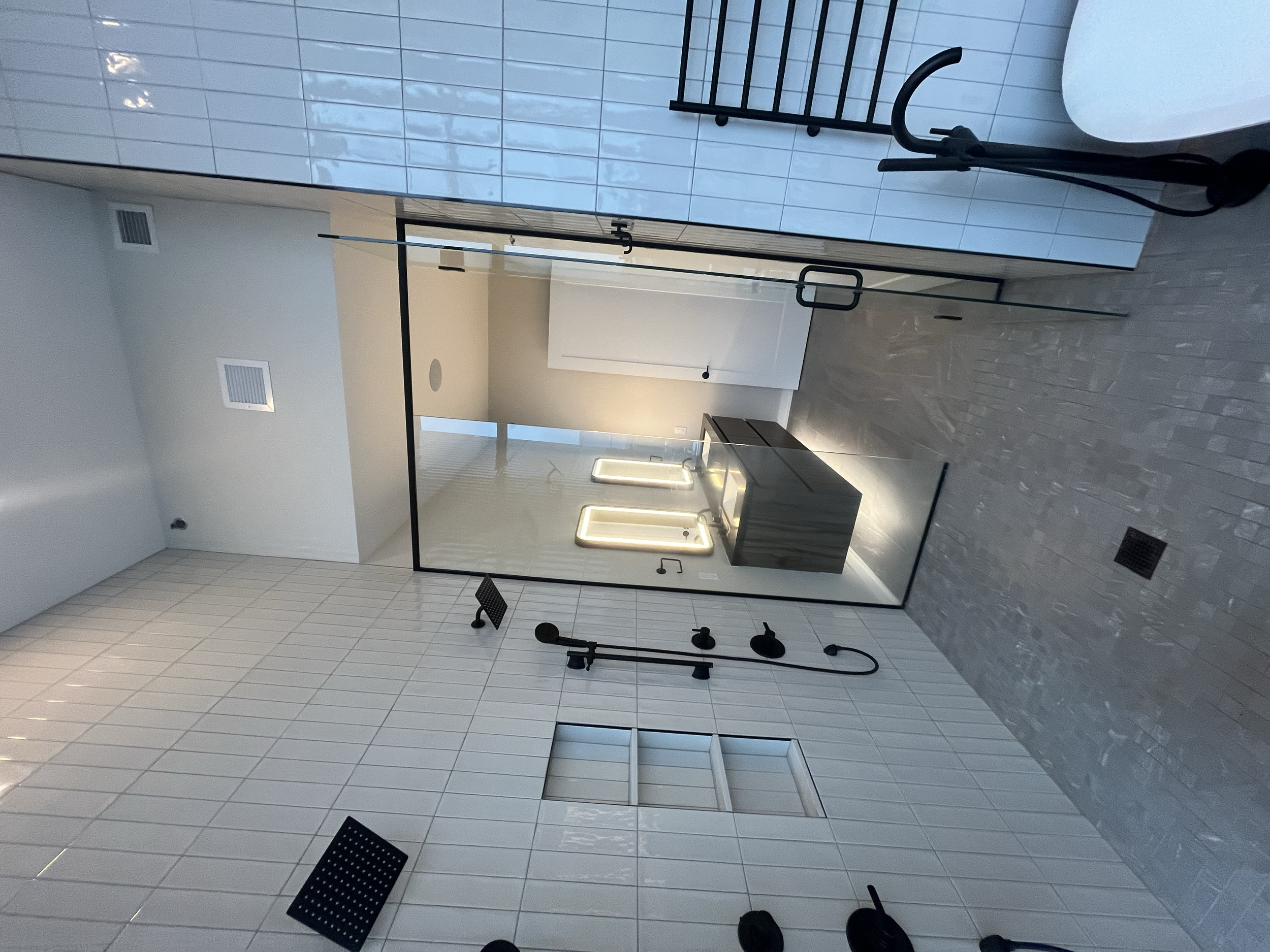 Modern bathroom with walk-in shower enclosed by glass, featuring black fixtures, a rain showerhead, and a black handheld shower. The space has white tiled walls, a gray tiled floor, and a large mirror reflecting a light-colored vanity and a window wi