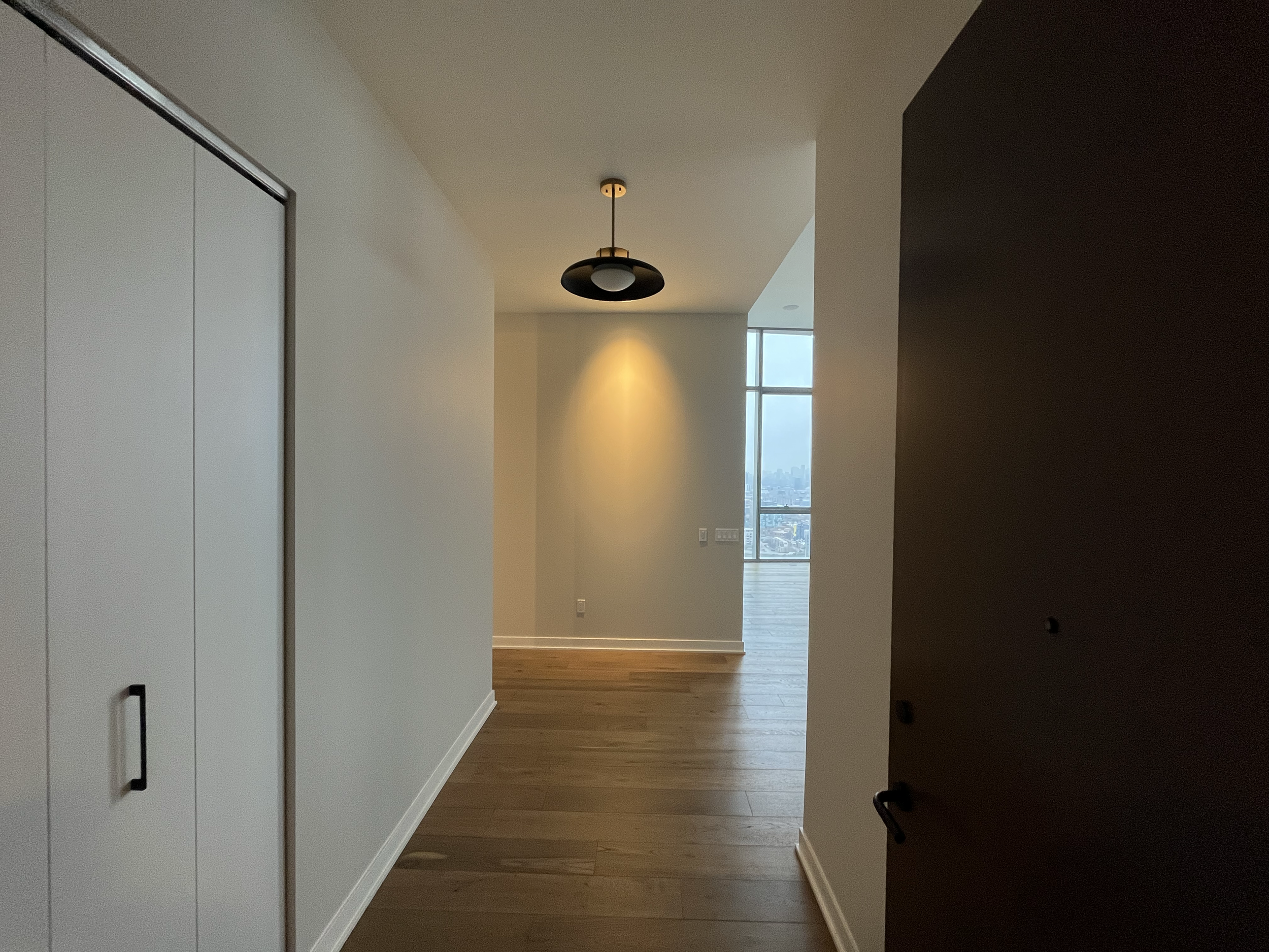 An empty modern apartment hallway with a wooden floor, white walls, a black ceiling light, and a large window showing a city skyline in the background.