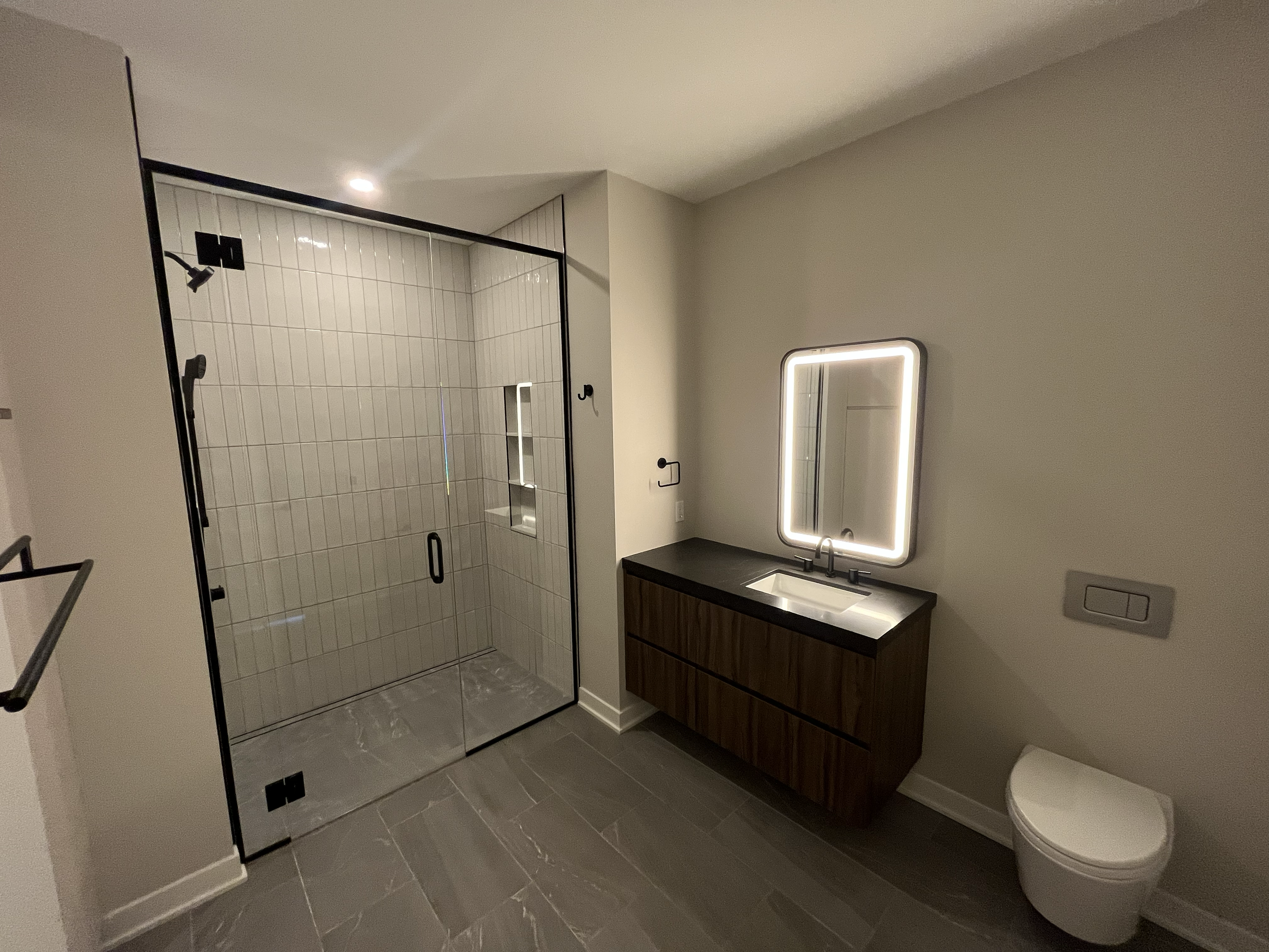 Modern bathroom featuring a glass-enclosed shower with dark framing, a wooden vanity with a black countertop and illuminated mirror, and a wall-mounted toilet.