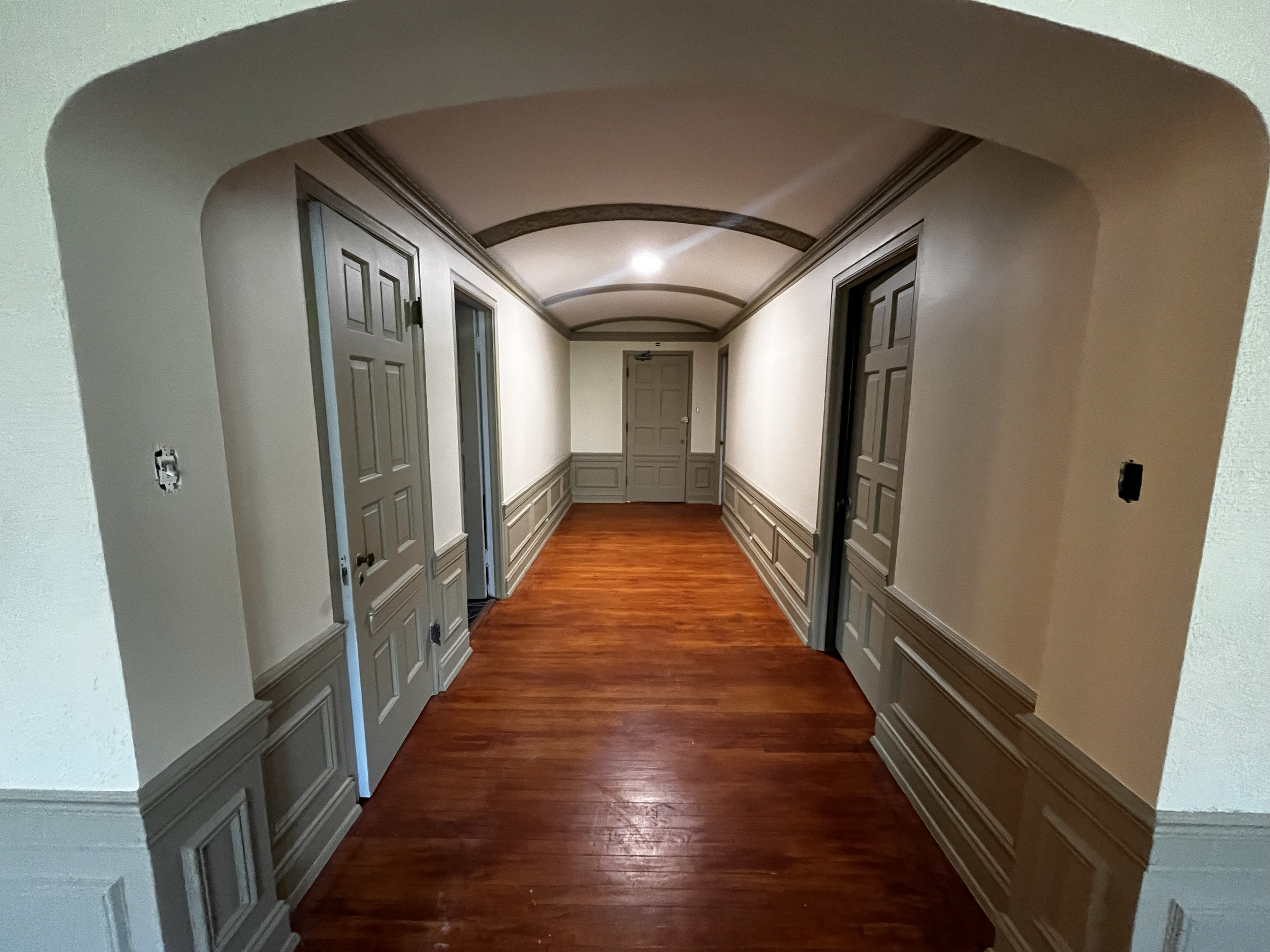 Empty hallway with cream-colored walls, wooden wainscoting, multiple closed doors, and a hardwood floor.