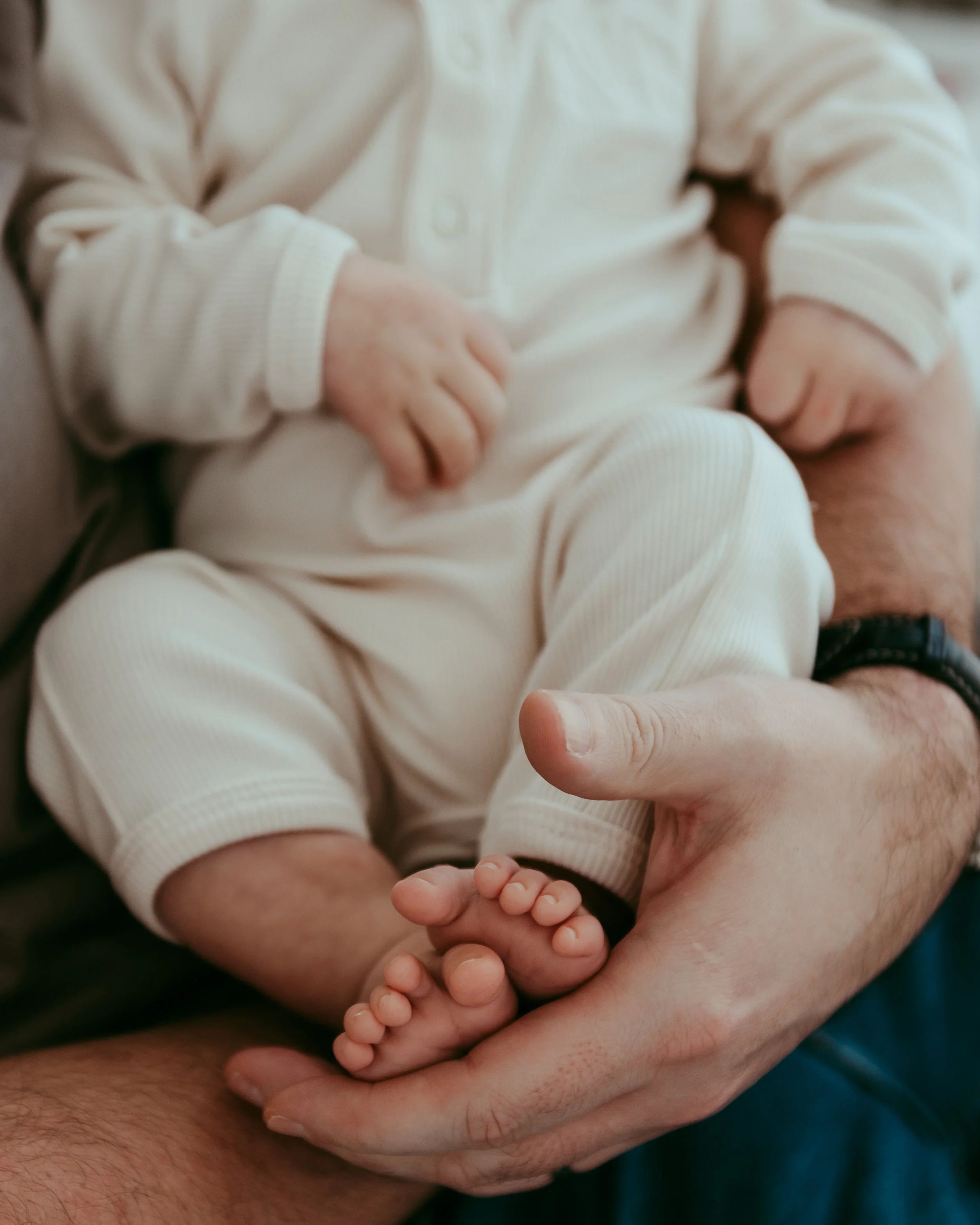 Close-up of a baby sitting on an adult's lap, with the adult holding the baby's feet in their hand. The baby is dressed in a cream-colored outfit.