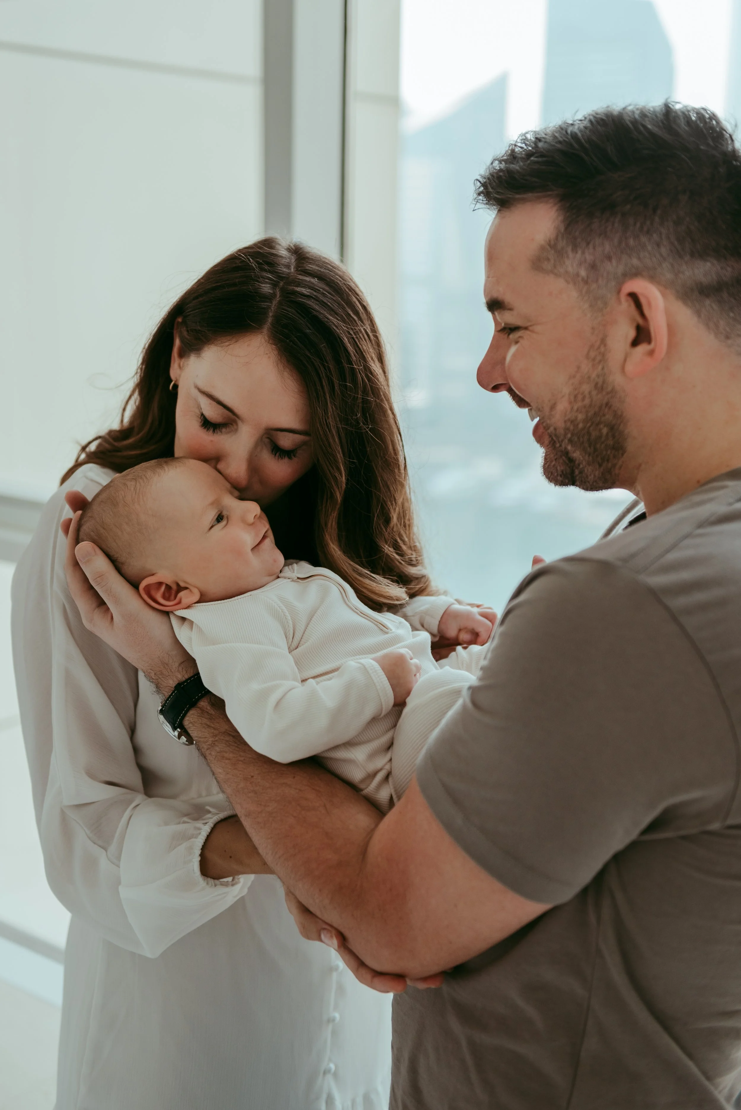 A woman and a man hold and look affectionately at a baby inside a modern house with large windows and cityscape in the background.
